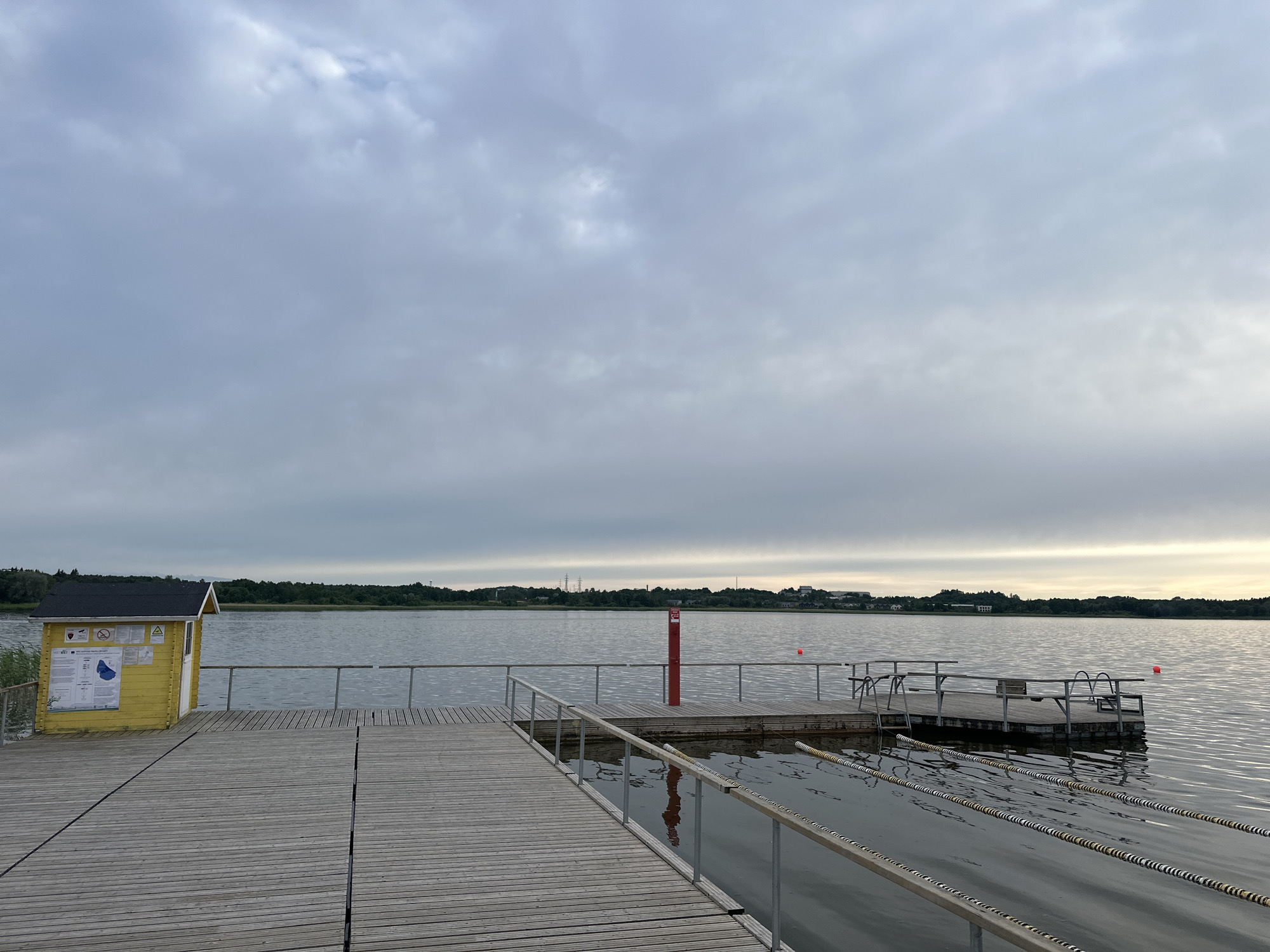 A wooden pier extends into a calm body of water under a cloudy sky. On the left side of the pier, a small yellow utility shed with a black roof has signs posted on its exterior. The pier is made of light-colored wooden planks and has metal railings. Further out into the water, a floating platform with metal railings and ladders is anchored. Several buoys are visible in the water, including a red one near the floating platform and another further to the right. In the distance, across the water, a treeline borders the horizon, with some buildings and industrial structures visible. The scene is an outdoor public space, likely a waterfront recreational area.