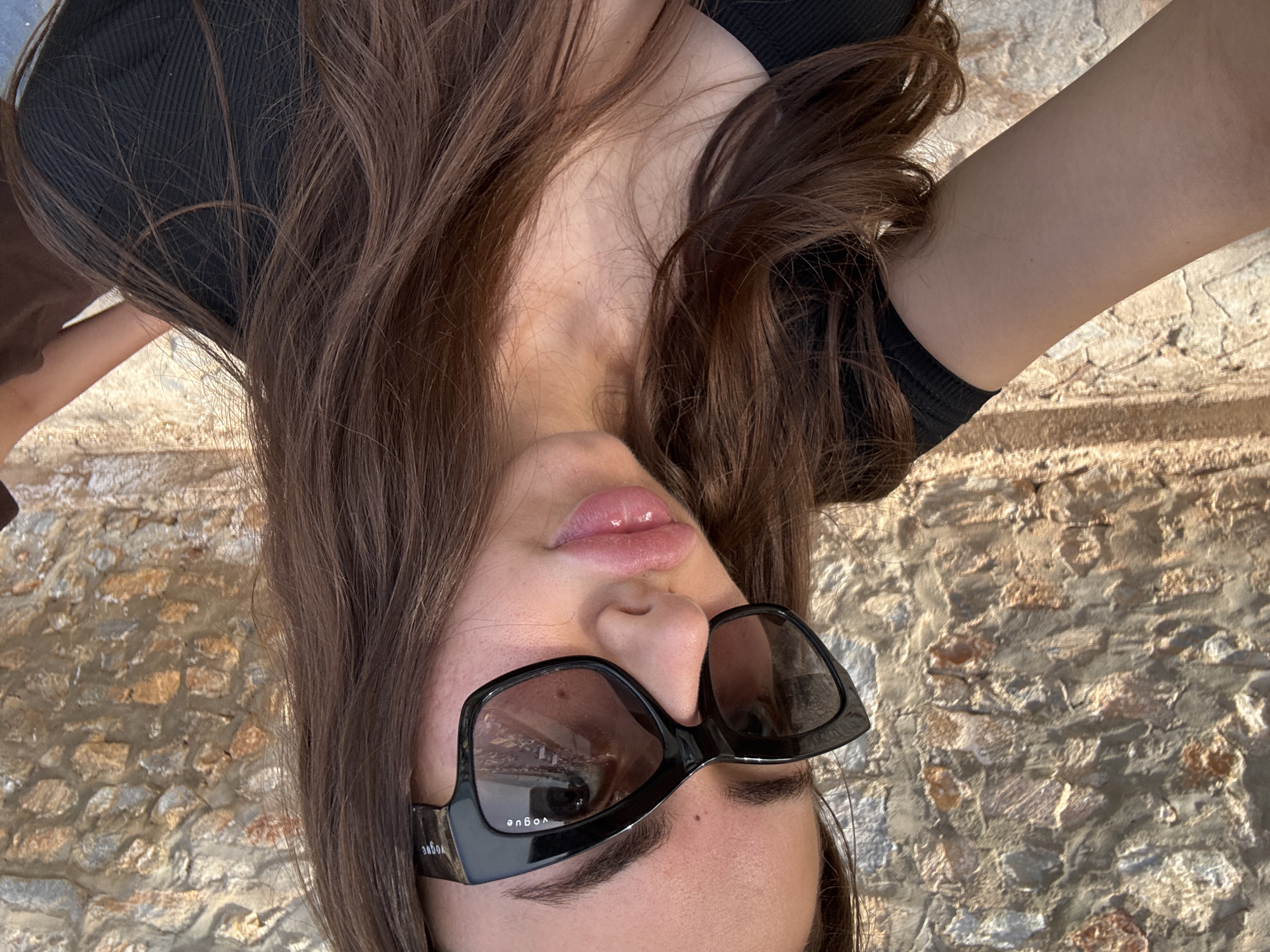 A young woman with long, brown hair and visible skin on her shoulders and chest poses for a selfie in front of a stone wall. She is wearing black, ribbed clothing and black sunglasses with "VOGUE" written on the temples. The sunglasses are reflective, showing a blurred image of an outdoor scene with some greenery and structures. Her lips are slightly parted and appear to be glossy. The background is a textured stone wall, appearing to be an exterior surface, possibly in Orihuela, Spain, given the architectural style of the stone work. The lighting suggests it is daytime, with a slight warmth to the illumination.