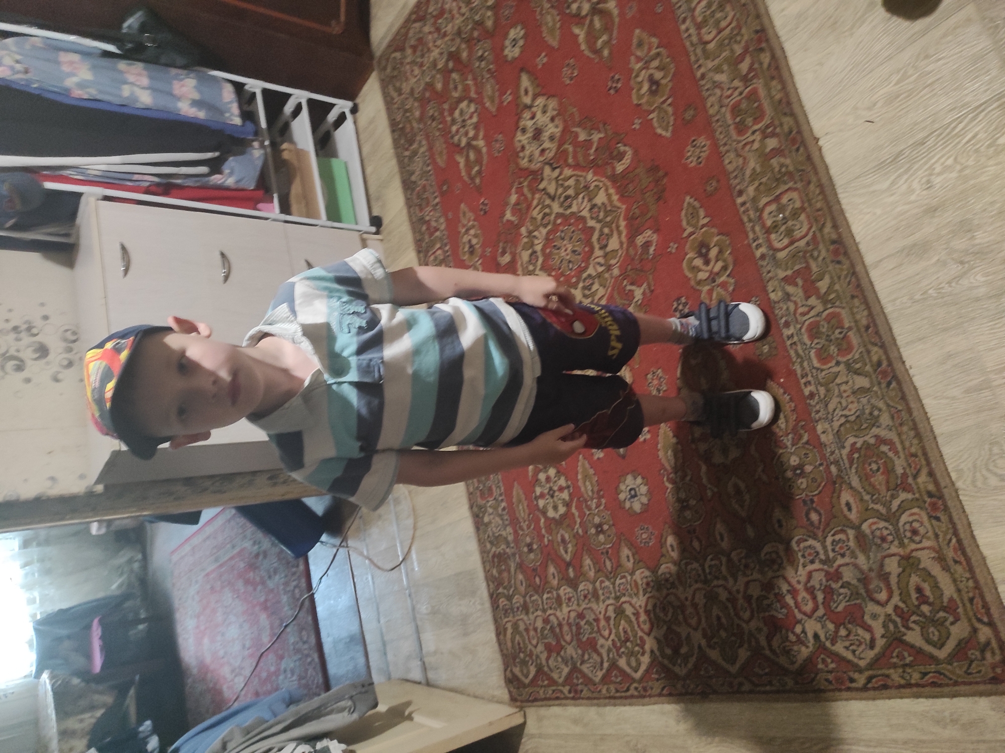 A young boy, appearing to be around 5-7 years old, stands on a red oriental rug in what appears to be a residential interior. He is wearing a horizontally striped polo shirt in shades of blue, grey, and white, dark blue shorts with a "Spider-Man" graphic, and black sneakers with white soles. He is also wearing a baseball cap with a colorful, abstract pattern. The boy is facing the camera and appears to be posing.
The background suggests a room with furniture, including a white dresser with drawers and a shoe rack with clothing and other items. A red rug is also visible in the background. The flooring appears to be wood laminate. The lighting is dim, and there is a shadow cast on the rug from the boy. The overall impression is a snapshot taken inside a home. The location is identified as the city of Kamianske, Ukraine.