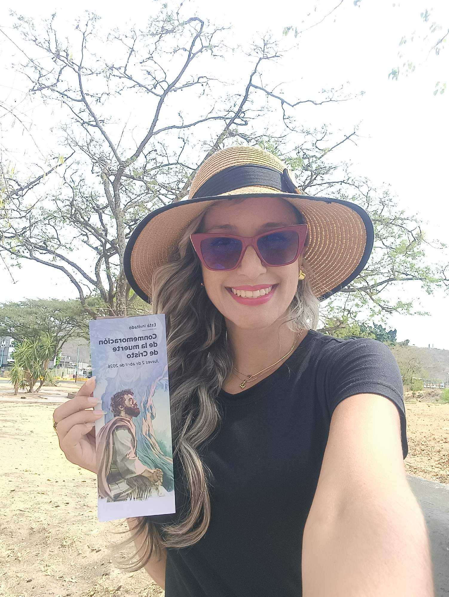 A female subject is captured in a front-facing, selfie-style photograph outdoors in La Victoria, Venezuela. She smiles widely, wearing a wide-brimmed straw hat with a dark band, red-framed cat-eye sunglasses with dark lenses, a black short-sleeved t-shirt, gold stud earrings, and a gold necklace with two small pendants. Her multi-toned, wavy hair extends over her shoulders. She holds a folded pamphlet in her left hand, facing the camera. The pamphlet displays mirrored text reading "Esta invitación," "Conmemoración de la muerte de Cristo," and "Jueves 2 de abril de 2020." An illustration on the pamphlet shows a kneeling, robed figure (Jesus Christ) under a tree, looking upwards. The background features a bright, likely sunny sky. A large, sparsely-leafed tree stands directly behind the subject. The ground is composed of light-colored earth or sand. To the left, several tall, palm-like plants are visible, along with distant roadside barriers, and indistinct structures. A dry, distant hill with sparse vegetation and vertical structures on its summit is visible to the far right in the background.