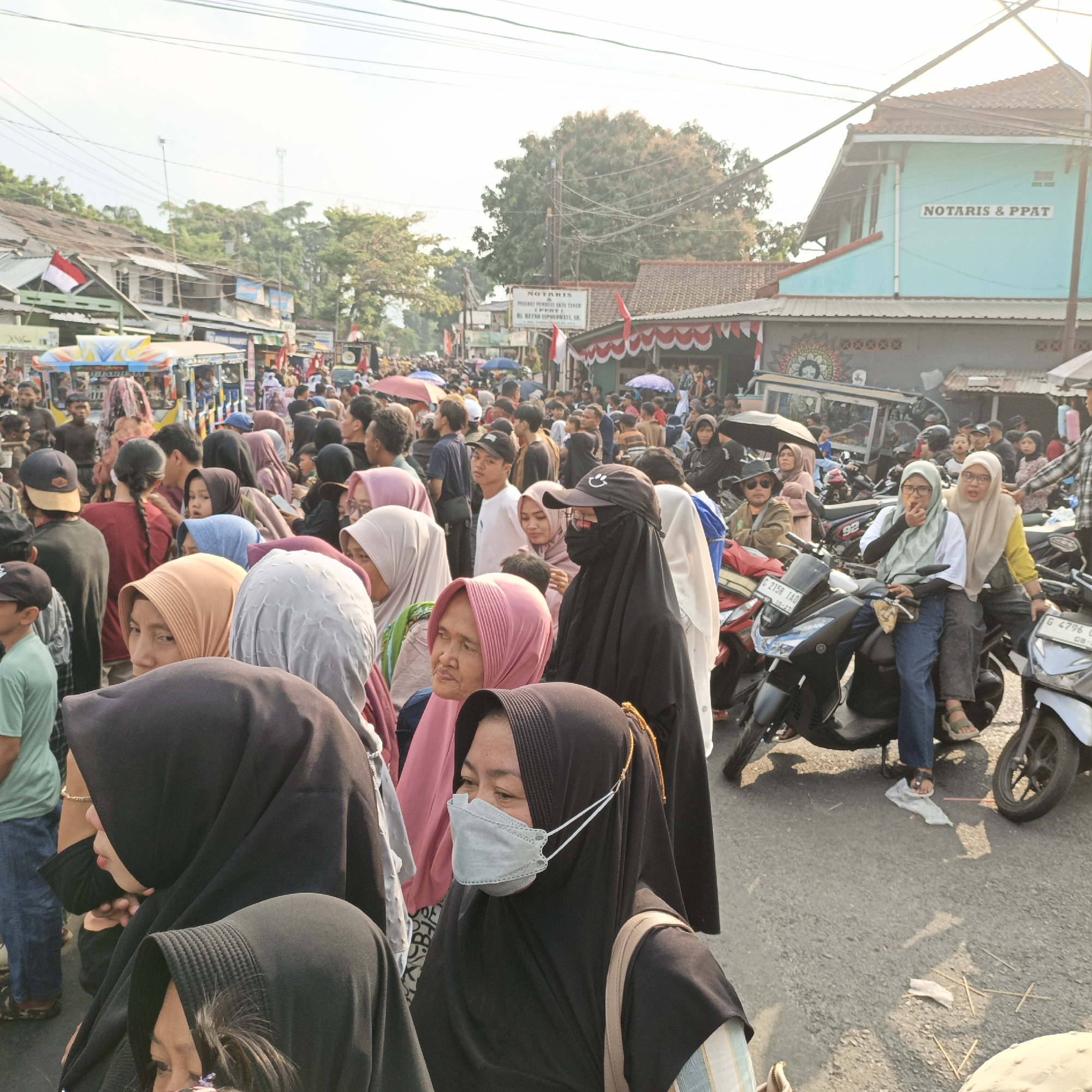 This is a medium-shot, eye-level, outdoor photograph taken on a street in Randudongkal, Indonesia, likely during a daytime event or gathering. The scene is crowded with people of various ages, many wearing headscarves (hijabs) and some wearing masks, indicating a public assembly. In the foreground, several individuals are prominently featured, their faces partially or fully visible. One woman in the immediate foreground is wearing a black hijab and a light blue face mask, looking to her left. To her left, another woman in a black hijab is visible. Behind them, a crowd stretches down the street, with some individuals on motorcycles parked on the roadside.
The background shows buildings lining the street, one of which is painted light blue and has a sign that reads "NOTARIS & PPAT". There are also tents, food stalls, and parked motorcycles, suggesting a market or community event. Red and white Indonesian flags are visible, adorning some of the buildings and stalls, which often signifies national celebrations or holidays. The overall impression is of a bustling, lively street scene in a populated area of Randudongkal. The lighting suggests it is a sunny day, with shadows cast on the asphalt road.