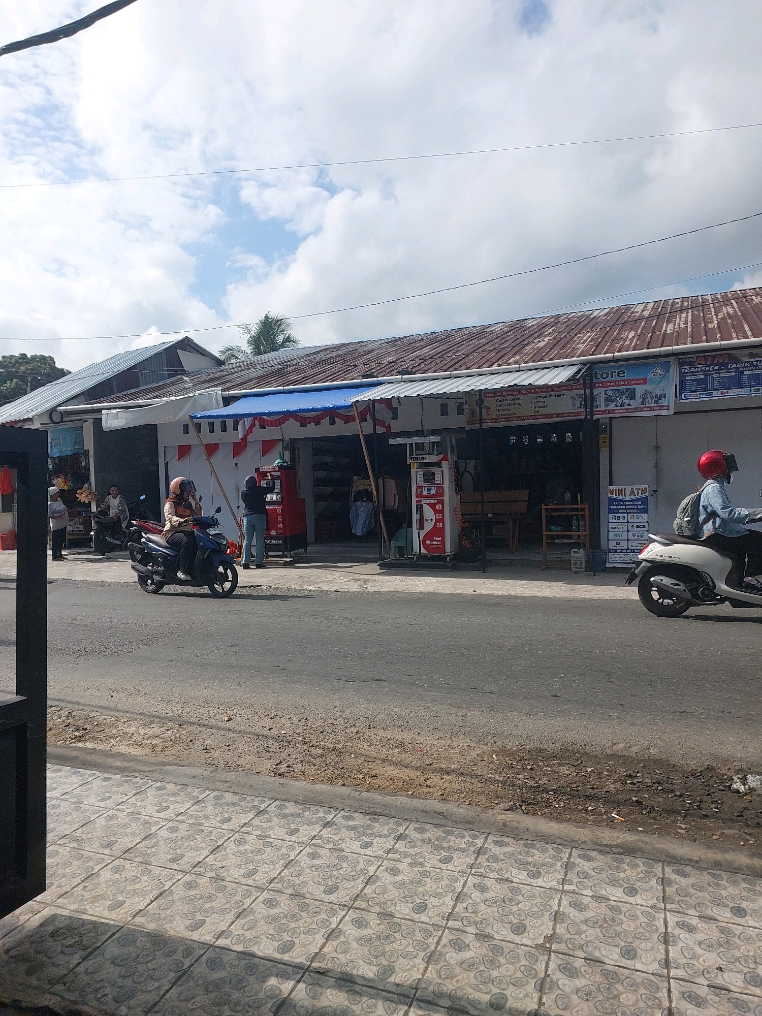 The image shows a street scene in Ambon, Indonesia, under a partly cloudy sky. In the foreground, a paved sidewalk with a stone pattern lines the edge of a road. On the road, a person wearing a red helmet rides a white scooter towards the right of the frame. Further down the road, a person in a beige hijab rides a dark blue scooter, also moving towards the right.
Adjacent to the road is a row of buildings with corrugated metal roofs. The buildings house various businesses, including a "store" selling clothing, indicated by a sign that reads "Herbal Berbagai Macam Pakaian Second Import," and a "Pertamini" fuel dispenser, a common type of small, independent gasoline station in Indonesia. Another sign above the store mentions "Celana Jeans," "Kaus Oblong," and "Blouse," all types of clothing. To the right of the store, a "MINI ATM" service is advertised, with logos for BRI, BNI, BCA, and Mandiri. Several people are visible near the establishments, some standing and some possibly entering or exiting. The overall impression is that of a bustling commercial area on a bright, overcast day.