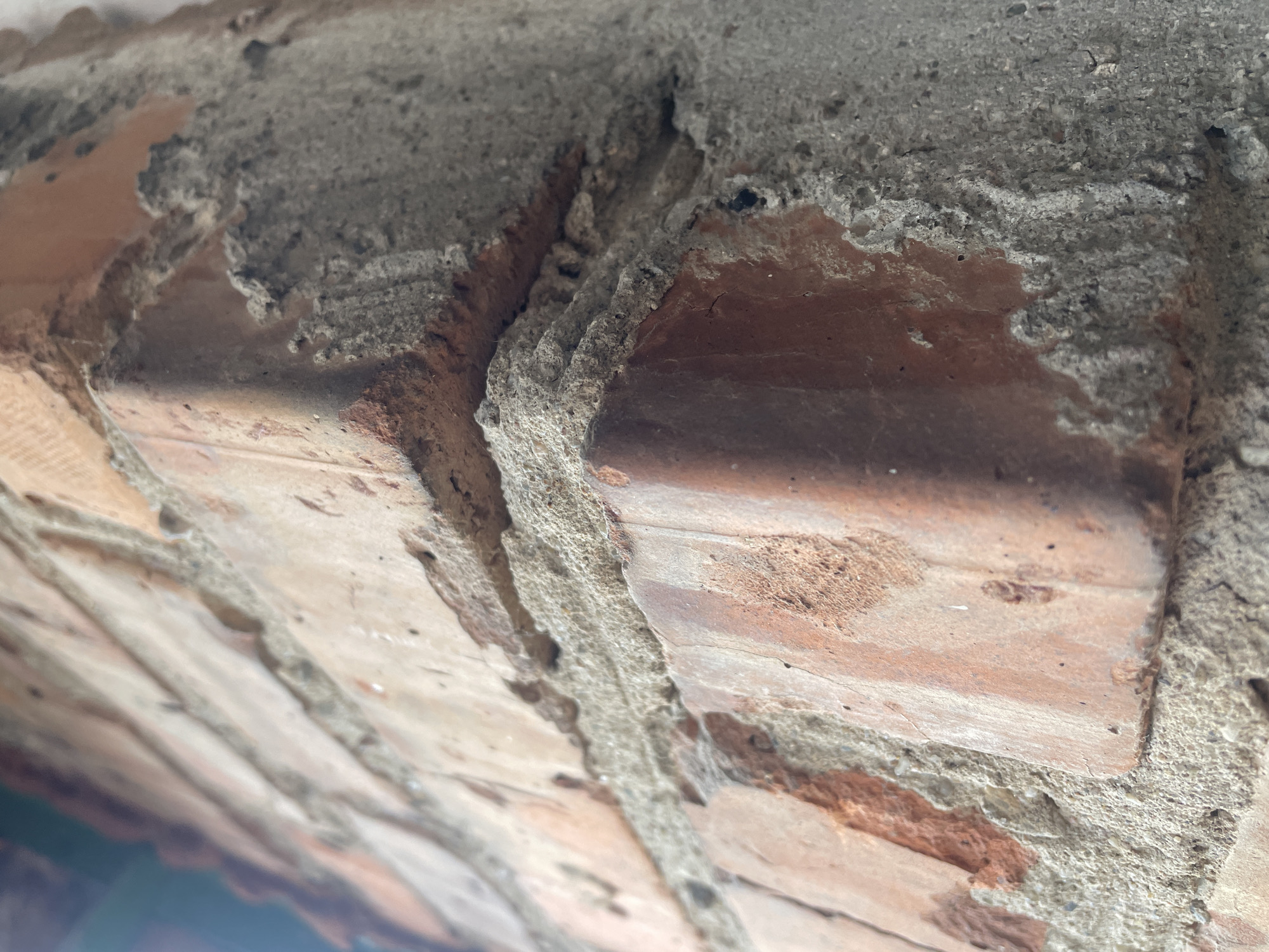 This is a close-up, low-angle, exterior photograph of the corner of a brick structure. The bricks are reddish-brown and have a rough, weathered texture, with visible mortar between them. The mortar appears to be a light gray, coarse material, also showing signs of wear and crumbling in places. The image is focused on the sharp corner where two walls meet, with the perspective leading the eye diagonally upwards and inwards. The lighting suggests it might be outdoors, possibly during the day, as there are some highlights on the brick surfaces. No people or animals are visible. The overall impression is one of aged construction.