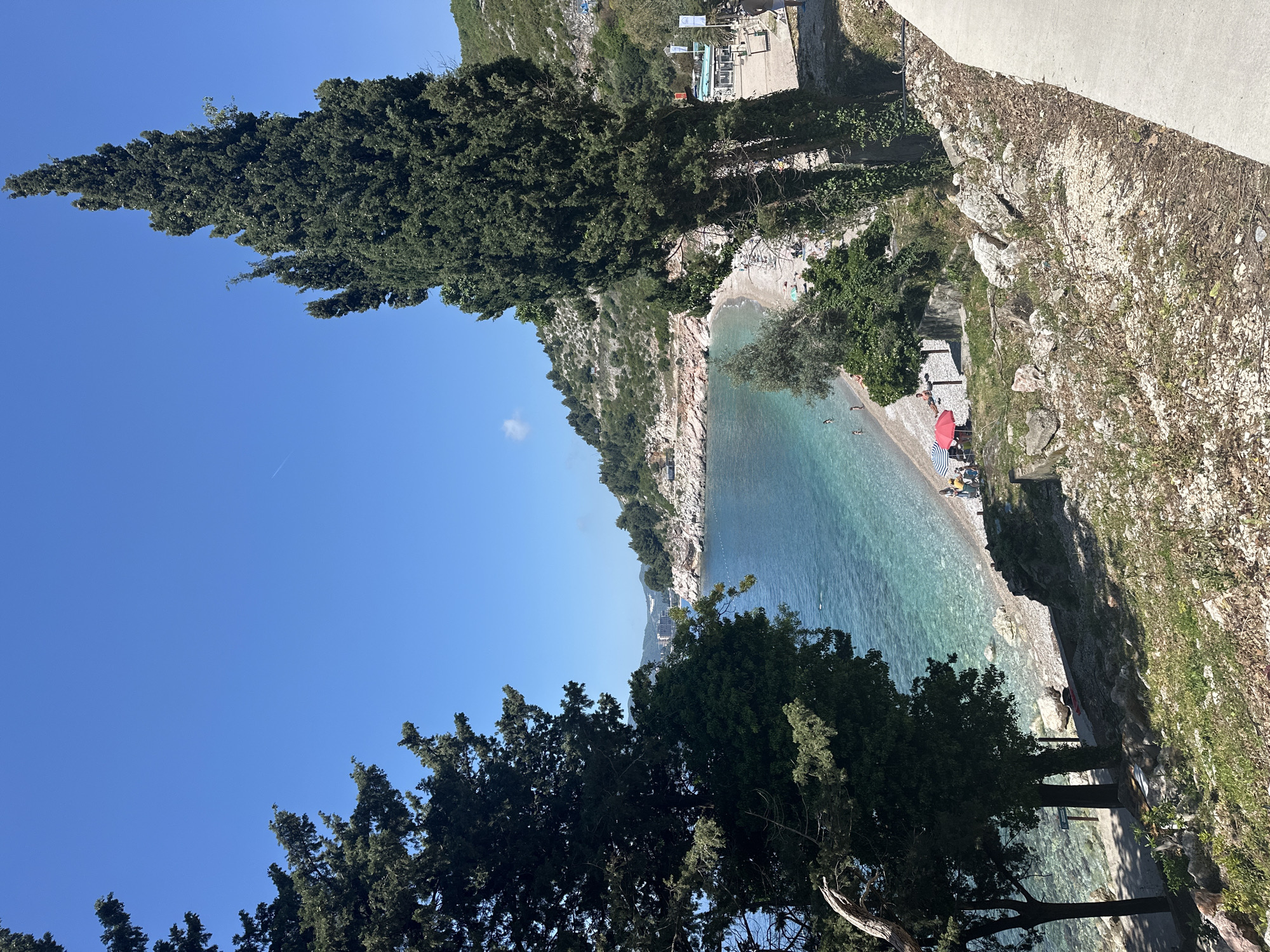 This image shows the beach in Kamenovo, Montenegro. The scene is dominated by a clear blue sky, large green trees, and turquoise water. People are sunbathing and swimming on the beach. Some objects, such as red umbrellas, are visible on the beach. The beach has rocky cliffs on one side. A concrete wall or path runs along the top of the cliff on the right side of the image.