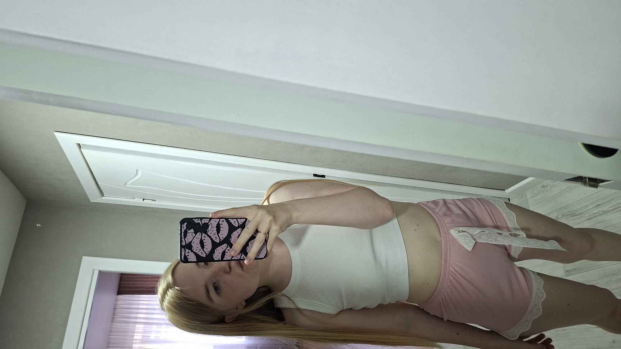 A young woman with long blonde hair is taking a mirror selfie in what appears to be a bedroom or dressing area. She is wearing a white ribbed tank top and pink shorts with white lace trim. She is holding a smartphone with a black case decorated with pink lips. The background shows a white door with decorative molding, a textured wall, and a light-colored wooden floor. The lighting suggests it is daytime. The overall impression is casual and intimate.