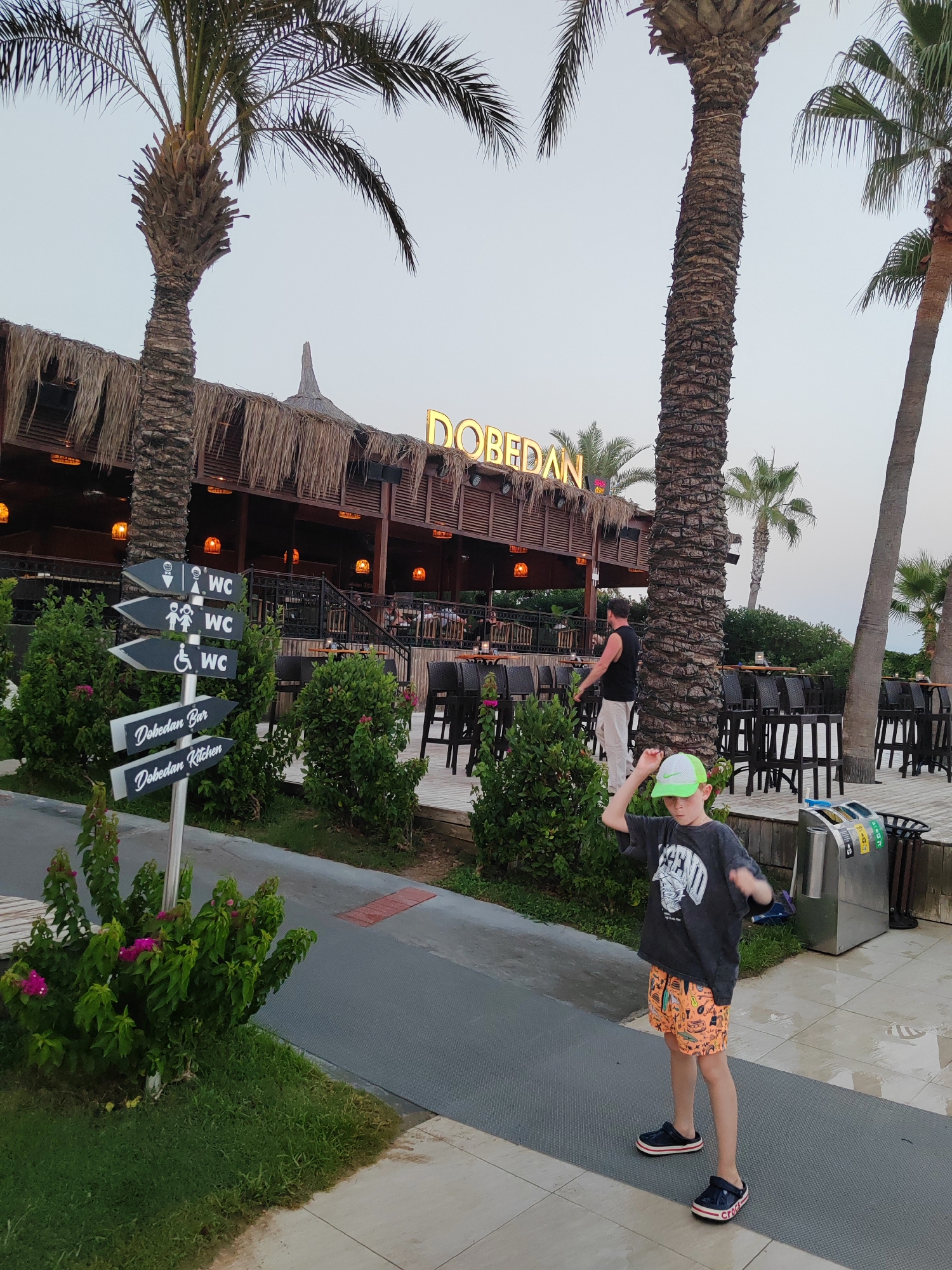 A young boy wearing a dark t-shirt, orange patterned shorts, a green cap, and dark sandals stands on a grey path in front of a building with a thatched roof. He is looking to his left and has his right arm raised. To his left, a signpost with multiple directional arrows indicates "WC," "Dobendan Bar," and "Dobendan Kitchen." The building appears to be a restaurant or bar, with tables and chairs visible outside. Palm trees are prominent in the foreground and background. The sky is overcast. The location is identified as Unknown, Unknown.