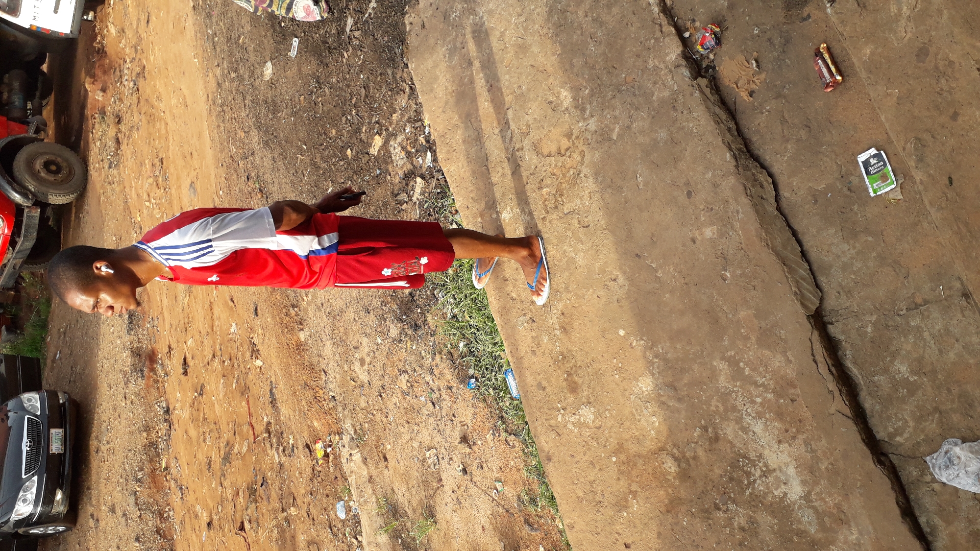 The image shows a man in Nkpor, Nigeria, standing on a cracked concrete surface. He is wearing a red and white short-sleeved shirt, red shorts, and blue and white flip-flops. He has an earpiece in his ear and appears to be looking at a mobile phone. The surroundings include a dirt ground, a black car, and the wheel of a red truck. Scattered litter is also visible on the concrete.