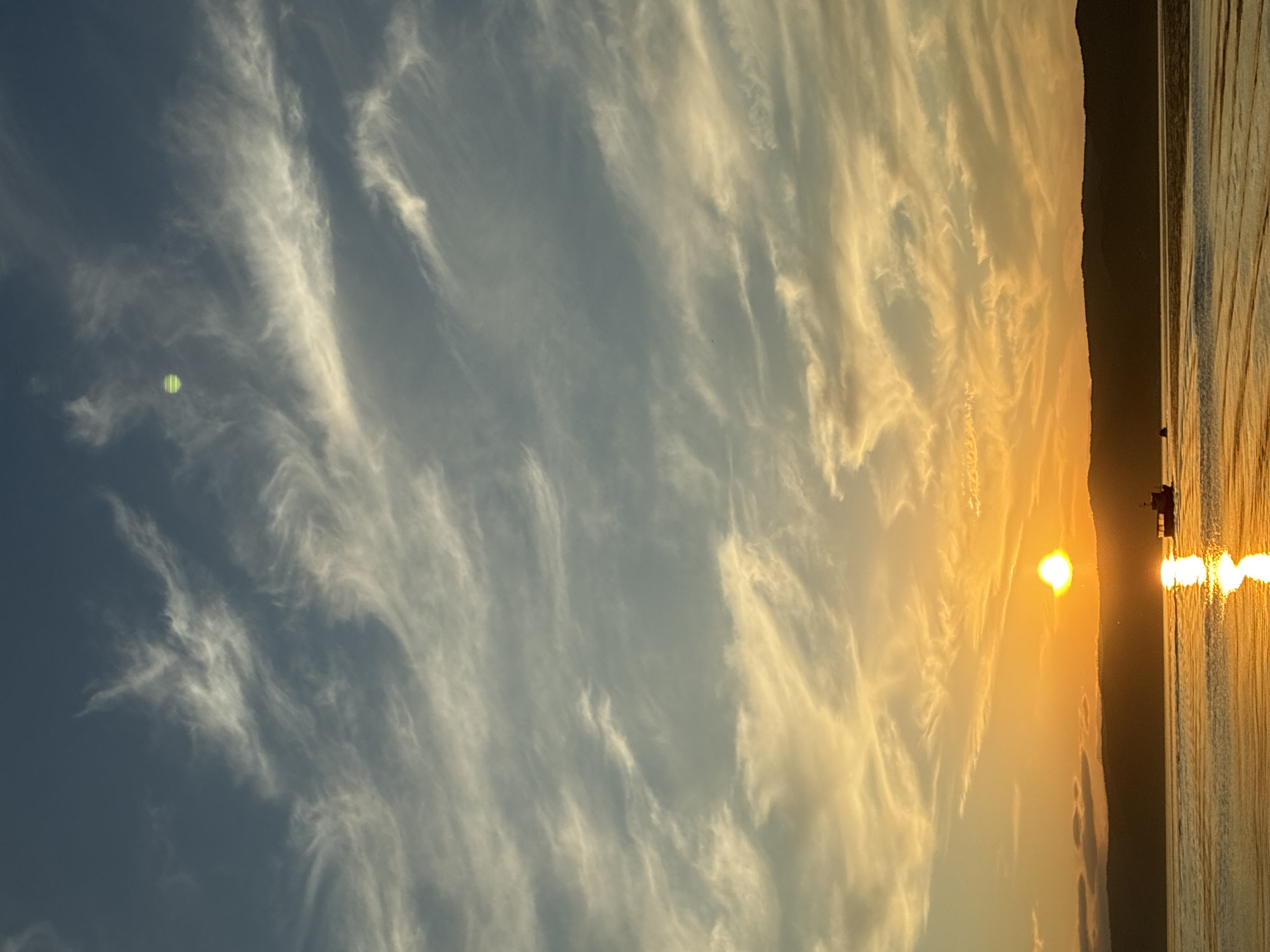 The image depicts a sunset over a body of water, likely Lake Baikal, near the settlement of Listvyanka, Russia. The sky is filled with wispy clouds, transitioning from a deep blue at the top to vibrant oranges and yellows near the horizon. The sun, a bright orange orb, is setting behind a silhouetted mountain range. The water reflects the golden hues of the sunset, creating a shimmering effect. In the foreground, the water is disturbed by the wake of a small boat, a dark silhouette against the bright water. Another smaller boat is visible in the distance. The overall scene is serene and captures the beauty of a natural landscape at dusk.