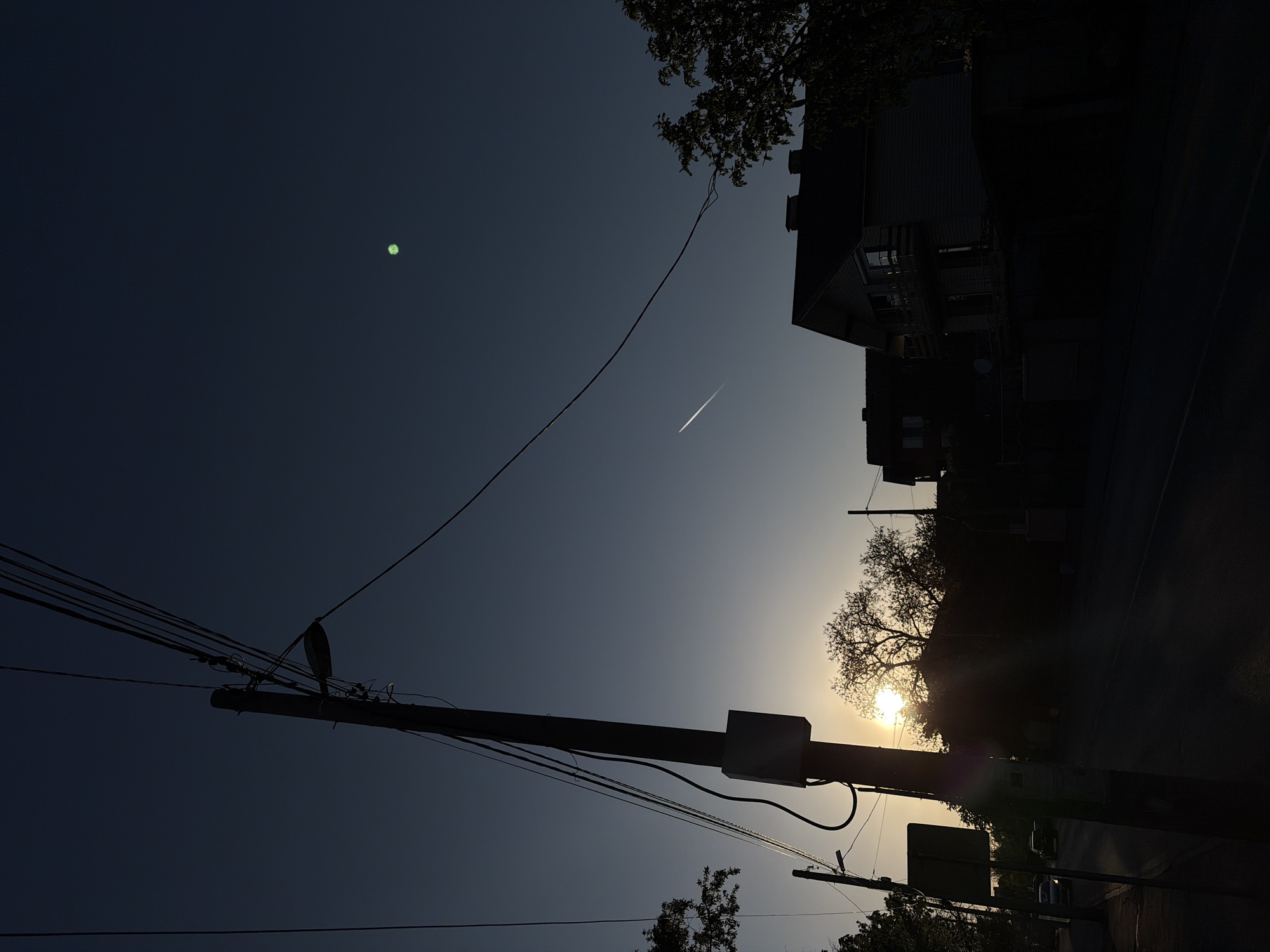 The image, captured in Sofia, Bulgaria, presents a low-light street scene at either sunrise or sunset, characterized by strong backlighting. The sky dominates the upper portion of the frame, transitioning from a dark blue-grey in the upper left to a brighter, warm golden hue towards the lower right where the sun is partially visible. The sun itself is a very bright and intense light source, creating significant lens flare and haze around it, and illuminating the outlines of nearby trees.
A prominent utility pole stands as a dark silhouette in the left-center foreground, extending vertically from the bottom of the frame. Numerous power lines and communication cables stretch diagonally and horizontally from this pole, creating intricate patterns against the sky. Further to the right and lower, another smaller utility pole and some street-level infrastructure, possibly including a street sign, are also visible in silhouette.
On the right side of the image, the dark, indistinct silhouettes of buildings and dense tree foliage rise against the brighter sky, their details obscured by the extreme backlighting. A dark road or street is visible along the bottom of the frame, with faint, silhouetted parked cars discernible.
Notable details include a distinct, bright green circular lens flare in the upper left area of the sky, and a thin white contrail, likely from an airplane, streaking across the middle of the sky. The overall atmosphere is quiet and contemplative, with the low sun creating dramatic contrasts and emphasizing the dark outlines of the urban environment against the luminous sky. No discernible text is visible in the image.