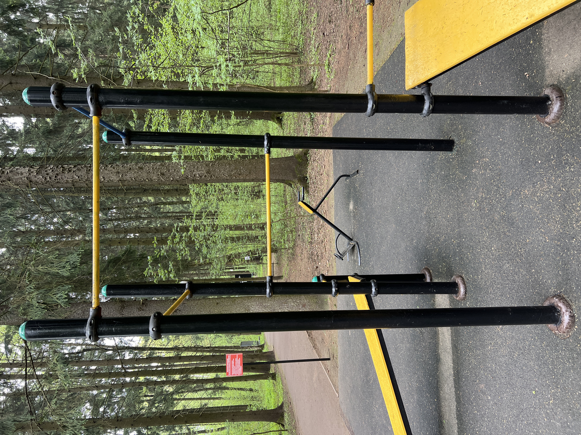 The image shows outdoor fitness equipment on a paved surface in Vnukovo, Russia. The equipment consists of black horizontal bars connected by yellow and blue vertical supports. A yellow platform is attached to the upper bar on the right side. The background features a forest with tall trees. A red sign is visible on the left side of the frame. The ground cover is asphalt or a similar material.