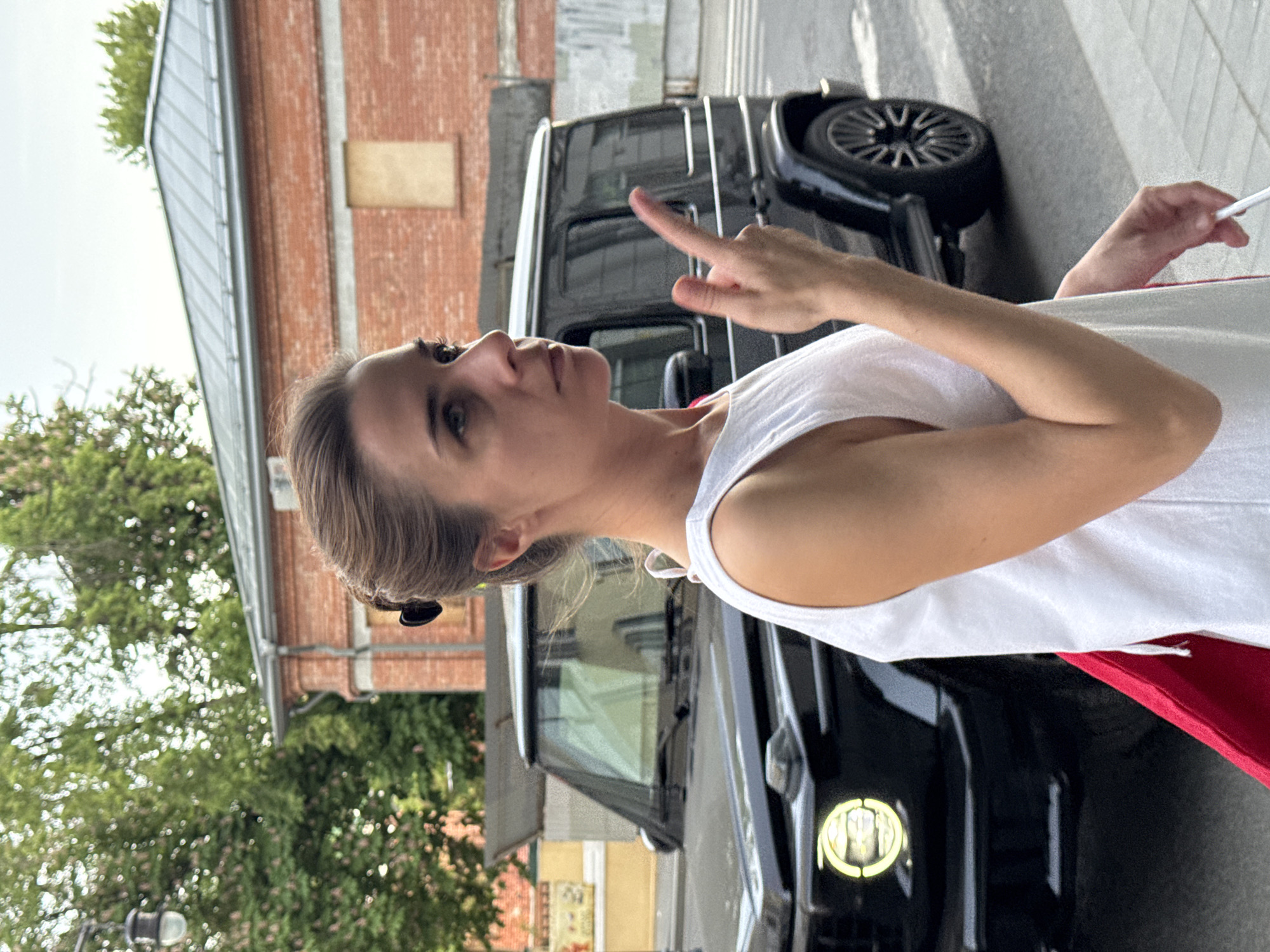 A woman with brown hair is standing in front of a black Mercedes-Benz G-Class SUV. She is wearing a white tank top and red pants. She is holding a cigarette in her right hand and pointing with her left hand. A brick building is in the background along with trees. The image was taken in Moscow, Russia.