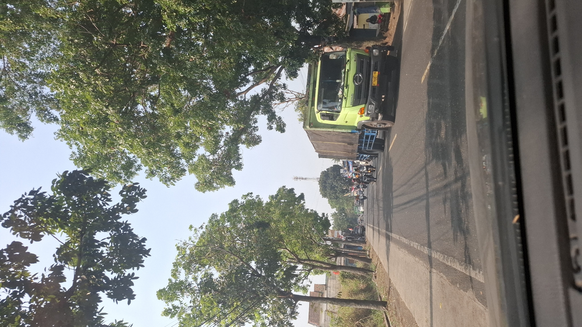 A bright, sunny daytime scene unfolds on a two-lane asphalt road in what appears to be the city of Ciwalen, Indonesia. The road is lined with mature trees casting dappled shadows on the pavement. On the left side of the road, a row of buildings, including houses with red-tiled roofs, are visible behind a line of trees and vegetation. On the right side of the road, a large, light green truck with a tarp-covered cargo bed is parked partially on the shoulder. Its license plate reads "D 9442 YB." A few individuals are seated on a bench under a large tree next to the parked truck. Several motorcycles and a few cars are traveling on the road, heading away from the viewer. The sky is clear and blue. The perspective suggests the image was taken from inside a vehicle.