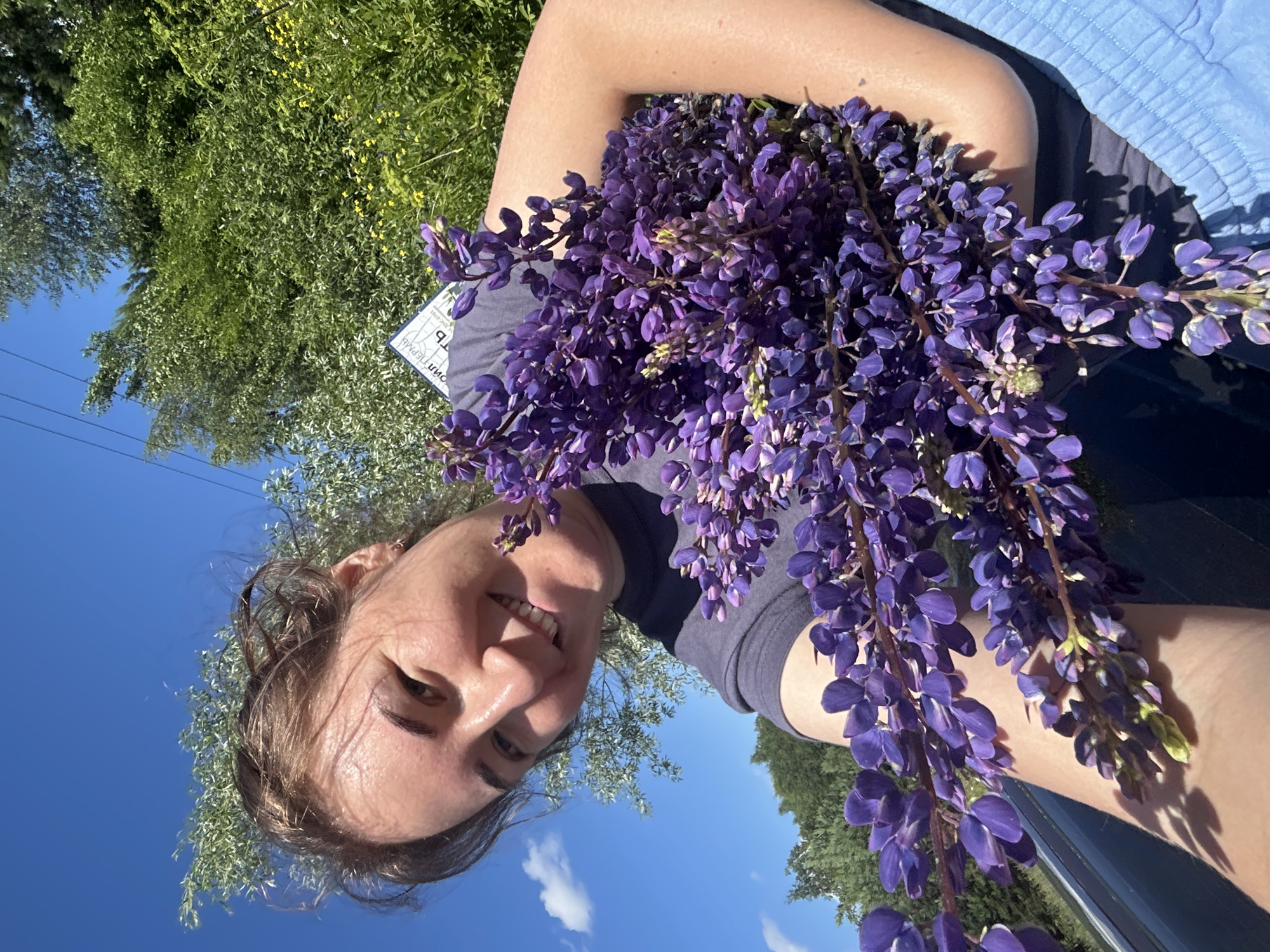 An outdoor, close-up, selfie-style photo shows a young woman with brown hair pulled up, smiling at the camera. She is holding a large bouquet of purple lupine flowers. The woman is wearing a dark blue t-shirt. Behind her, the background consists of lush green trees, a bright blue sky with a few white clouds, and a sliver of a gray car. A sign with Cyrillic text is visible in the background, slightly out of focus. The overall lighting suggests a sunny day. The setting appears to be a natural environment, possibly a roadside in Верх-Рождество, Russia.