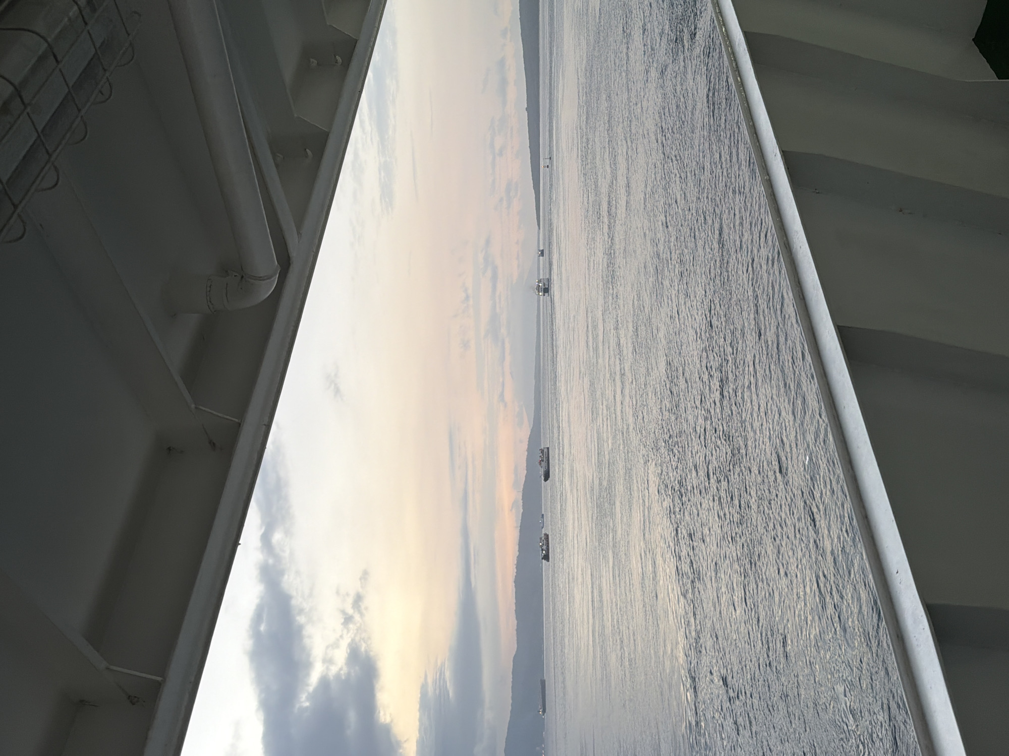 The image depicts a seascape viewed from the deck of a vessel. The perspective is angled, with the foreground dominated by the white structures of the ship, including pipes and beams. Beyond the ship's railings, a wide expanse of water stretches towards a distant, silhouetted landmass, likely a coastline. The water is textured with ripples, reflecting the soft, diffused light of dawn or dusk. Several small boats are scattered across the water, some appearing as dark silhouettes against the brighter sky, others with faint lights.
The sky transitions from a pale, luminous yellow-orange near the horizon to a soft blue with scattered, wispy clouds. The overall atmosphere is tranquil and serene, suggesting a peaceful moment at sea. The time of day is either early morning or late evening, given the low angle of the sun and the soft lighting. The location is identified as Melaya, Indonesia, implying a tropical or subtropical setting. No people are clearly visible in the image. The scene evokes a sense of calm and openness, with the vastness of the ocean and sky dominating the view. There is no visible text in the image.