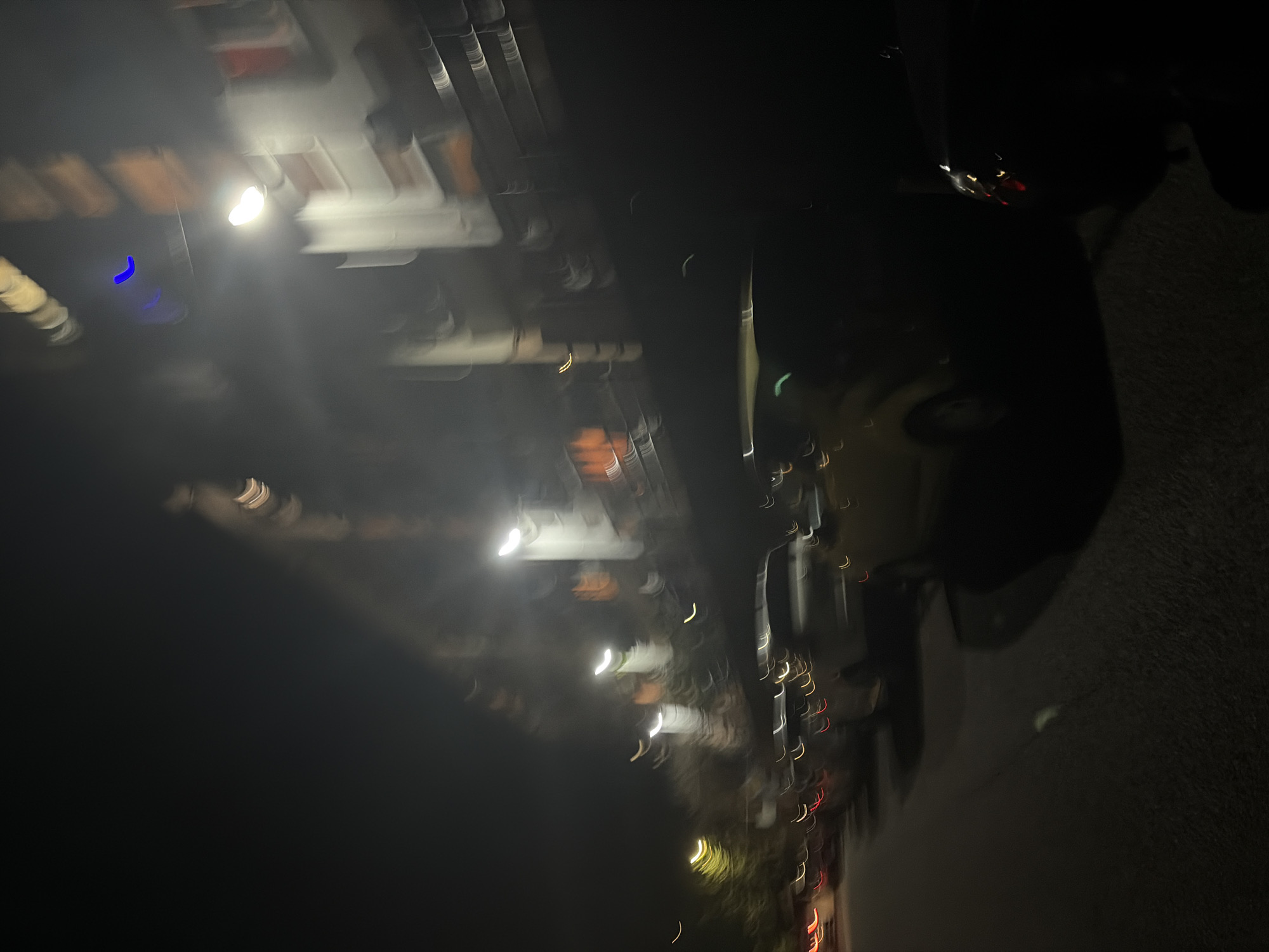 The media file is a heavily blurred and rotated photograph taken at night, likely depicting a street scene in Sochi, Russia. The primary subjects are numerous bright, streaky artificial lights, appearing mostly white, yellow, and orange, suggesting streetlights or lights emanating from buildings. These lights are distributed across the middle and upper-left sections of the image (when viewed upright). One distinct blue light streak is visible in the upper-left area.
The intense blur creates elongated light trails and soft glows, indicating significant motion during exposure or camera shake. Beneath and behind the blurred lights, indistinct shapes suggest buildings or structures lining a thoroughfare. A dark, textured area, possibly a road or ground surface, occupies the lower right and bottom portions of the frame.
The activity captured is the visual effect of motion blur on a night scene. The time of day is clearly night, characterized by darkness and artificial illumination. No specific details, such as architectural styles, vehicle types, individuals, or weather conditions, are clearly discernible due to the extreme blur. There is no visible text. The overall impression is an abstract night cityscape captured with significant motion.