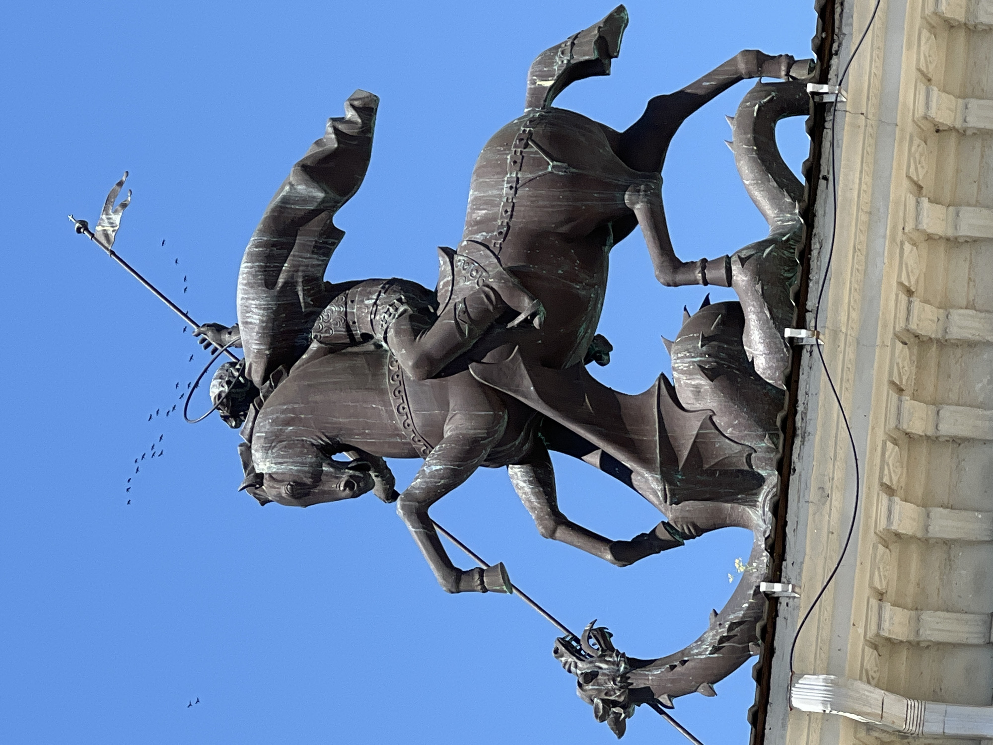 The image depicts a bronze statue of Saint George on horseback, slaying a dragon, atop a building in Krasnodar, Russia. The statue is dark bronze with green patina, indicating age and exposure to the elements. Saint George is depicted in armor, wearing a cape that billows behind him, and holding a spear that pierces the dragon. The dragon is coiled beneath the horse's hooves, with its head raised and mouth open in a snarl. The background is a clear blue sky, with a flock of birds in flight in the upper left quadrant of the image and a few more near a banner held by Saint George. The building below the statue appears to be made of stone and features architectural details like columns and decorative moldings. The statue is mounted on what looks like a roofline, with a gutter system visible below.
