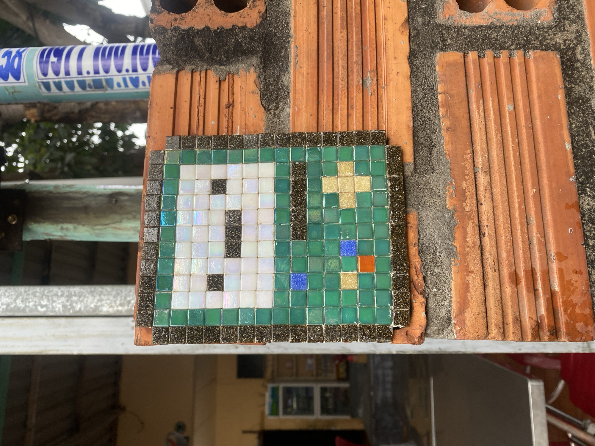 A mosaic tile depicting a stylized 'P' shape, predominately white on the left and gold on the right, set against a green background, is affixed to a weathered brick structure. The mosaic tile has a dark border. The location is indicated as Phu Thuy Ward, Vietnam, based on the presence of a Vietnamese advertising sign in the background and the style of construction. The surrounding environment appears to be an outdoor space, evidenced by the presence of foliage in the background and the lighting conditions.