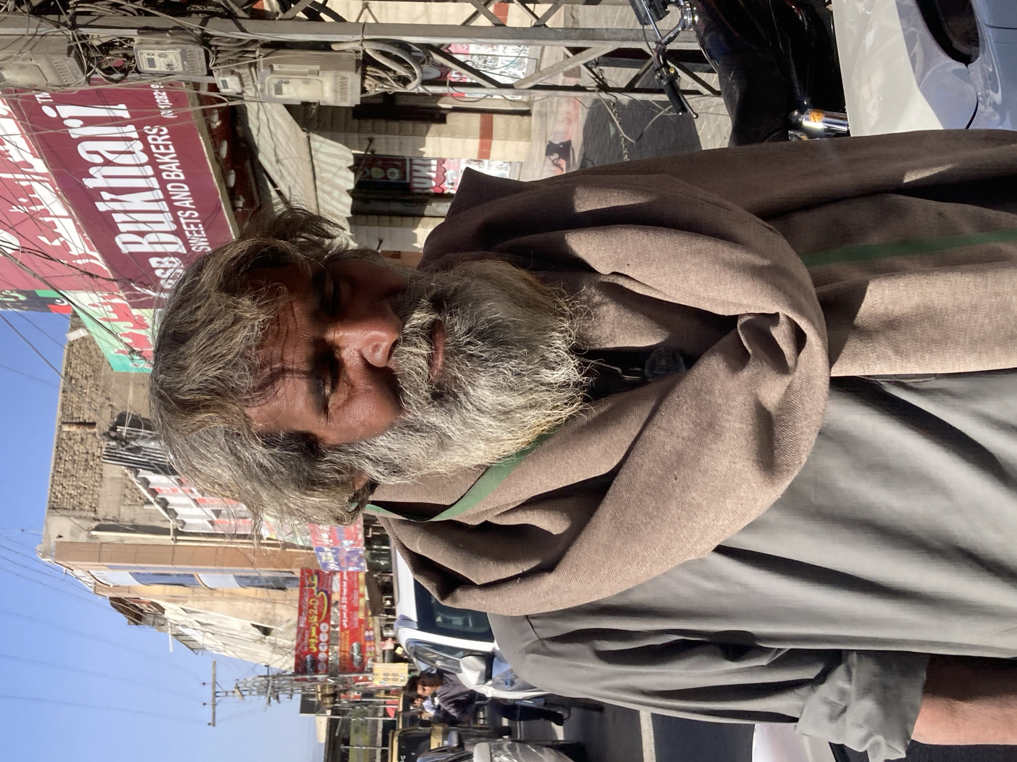 The image captures a man with grey hair and a beard, draped in a brown shawl over a grey shirt, seemingly in an urban area. Behind him, a street scene unfolds, revealing a sign in Urdu for "SB Bukhary Sweets and Bakers." Buildings line the street, and utility wires crisscross overhead. The presence of vehicles, including a motorcycle, suggests a bustling environment. The photograph appears to have been taken at street level. The location is in Quetta City Tehsil, Pakistan.
