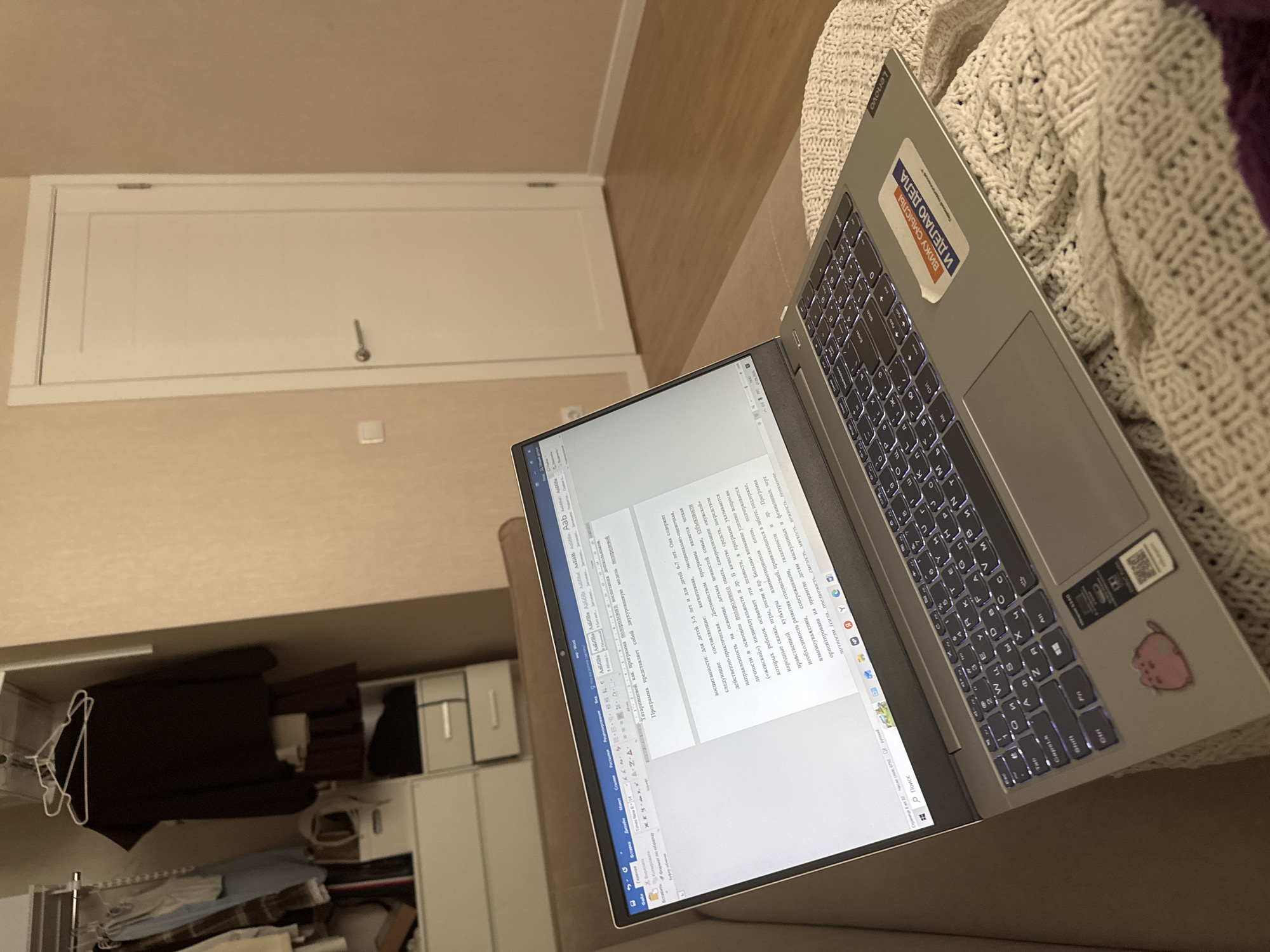 The image depicts an indoor scene, likely a residential interior, within the city of Perm, Russia. The perspective is from a slightly elevated position looking down towards a laptop situated on a seating area.
In the foreground and center, a silver-grey Lenovo laptop is open and rests on a cream-colored, textured knitted blanket or throw. The blanket covers a light brown or beige sofa or bed. The laptop screen displays a Microsoft Word document written in Russian, with text centered on the page. The document appears to discuss educational concepts, including "воспитании" (upbringing) and "программа полоролевого воспитания" (gender-role education program). The laptop's keyboard is backlit, and its layout is consistent with a Russian keyboard. On the laptop's palm rest, there are three stickers: a pink cartoon cat-like character on the left, a small black and white sticker (possibly a QR code or product label) to its right, and a larger rectangular sticker on the far right that reads "ВИЖУ СМЫСЛЫ И ДЕЛАЮ ДЕЛА" (I see meanings and do deeds) in blue and red text on a white background. The "Lenovo" branding is visible in reverse orientation on the edge of the laptop chassis near this sticker, indicating the laptop is angled. A small segment of a dark purple fabric is visible at the bottom right corner of the frame.
To the left of the laptop, a portion of a built-in wardrobe or closet system is visible. It features white modular storage units with silver handles and open sections. Hanging clothes are visible, including a black long-sleeved top on a white hanger, a light blue shirt, and a plaid shirt. Other clothing items, such as a dark brown pleated skirt and a white item, are folded or stored on shelves within the unit.
In the background, to the right of the laptop, a closed white interior door with a silver handle is prominent. The door has subtle horizontal decorative grooves. A white rectangular light switch or outlet cover is visible on the wall to the left of the door. The walls throughout the visible room are painted in a light beige or tan color with a subtle textured pattern. The floor in the background, extending from the door, is light-toned wood laminate or hardwood.
No individuals are present in the image. The scene suggests a quiet, organized domestic environment.
