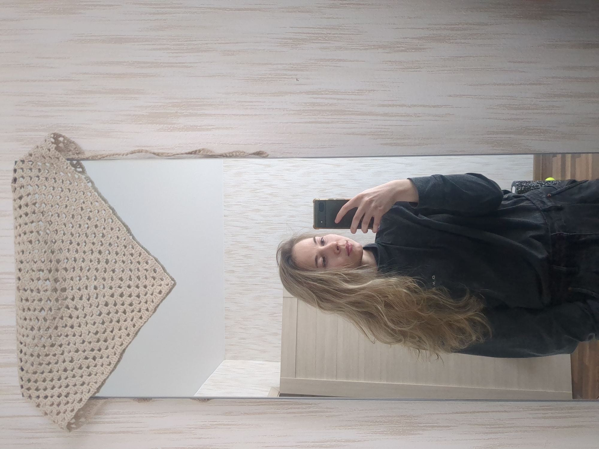 The image shows a reflection in a mirror, capturing a woman taking a selfie with her phone. The woman has long, blonde hair and is wearing a dark-colored outfit. A crocheted shawl is draped over a part of the mirror. The background appears to be an interior setting with striped wallpaper and a door in the reflection. There is no indication of the location being Saint Petersburg, Russia.