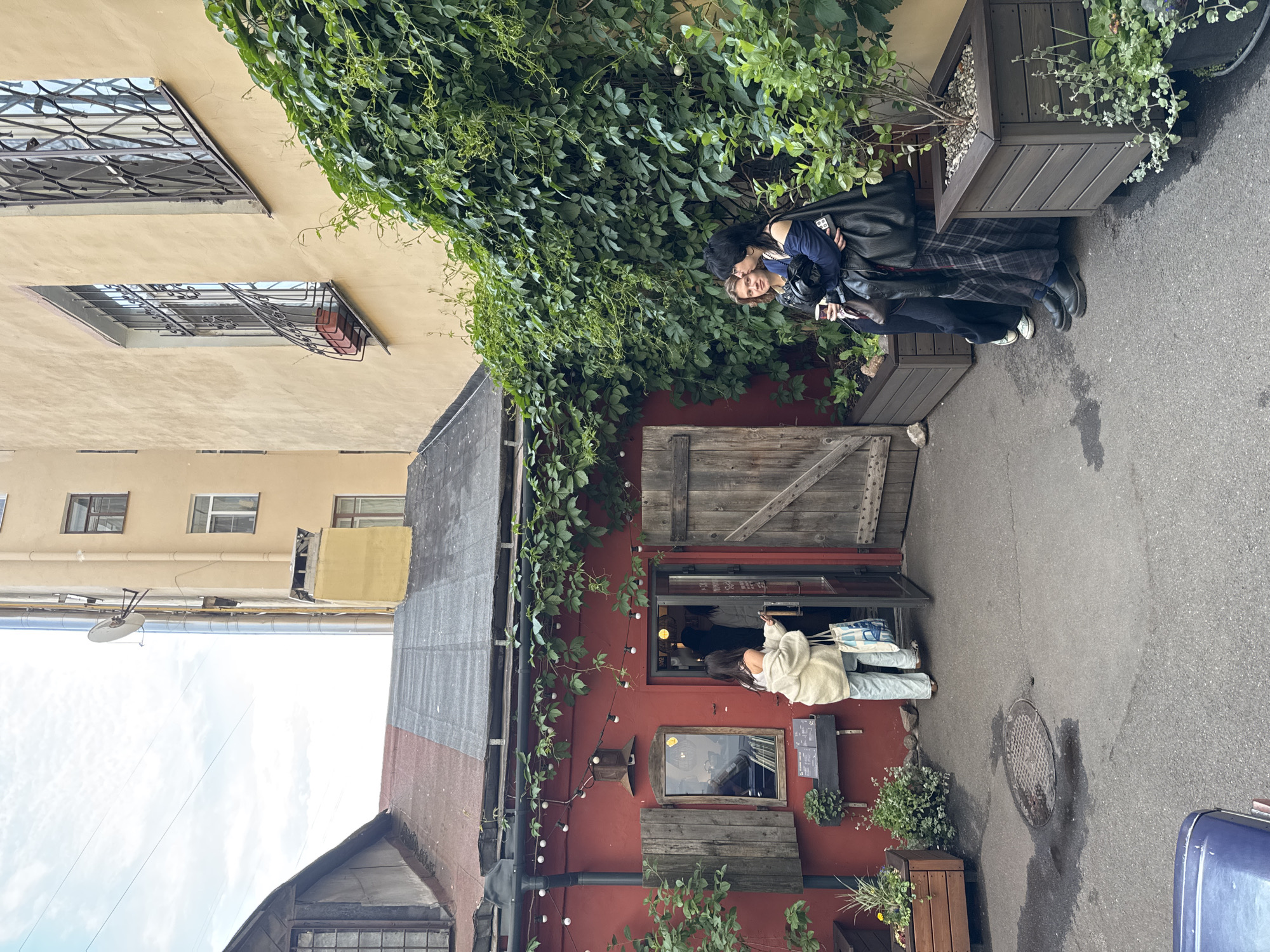 The image captures a narrow, cobblestone courtyard in Saint Petersburg, Russia, with a distinctive bohemian ambiance. On the left, a striking red building with a dark, sloped roof hosts an entranceway adorned with fairy lights and a wooden door. To the right of the entrance, a rustic wooden shutter is mounted on the red facade. Adjacent to the red building, a taller, cream-colored structure rises, featuring several windows with ornate metal grates. Lush green ivy cascades down the facade of the cream building, creating a natural canopy over a section of the courtyard. In front of the red building, a woman in a light-colored cardigan and light-colored pants, carrying a patterned tote bag, is entering the building. On the right side of the courtyard, two women stand near planters filled with greenery. The woman on the left, with blonde hair, holds a disposable coffee cup, and the woman on the right, with dark hair, wears a dark blue top and a plaid skirt, holding a black bag over her shoulder. The overall scene suggests a hidden gem, possibly a cafe or shop, tucked away in a courtyard.