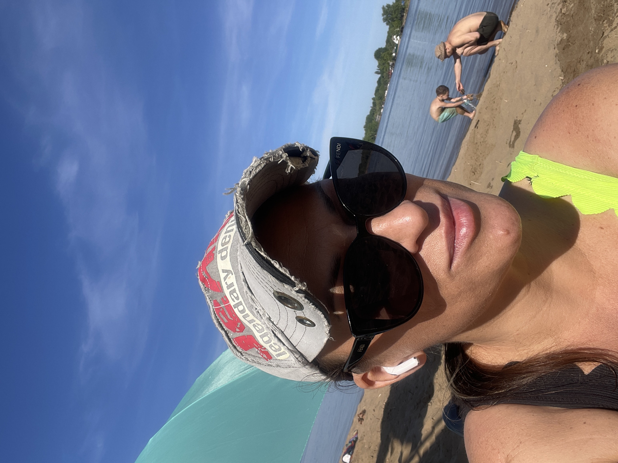 A selfie of a woman at the beach, wearing a weathered baseball cap, black Fendi sunglasses, and a bright green bikini top. An AirPod is visible in her ear. In the background, two figures are on the sand near the water's edge: an adult crouching and a child playing in the water. The environment appears to be a beach scene with water, sand, and vegetation on the opposite shore. The location is inferred as a beach near Saratov, Russia.