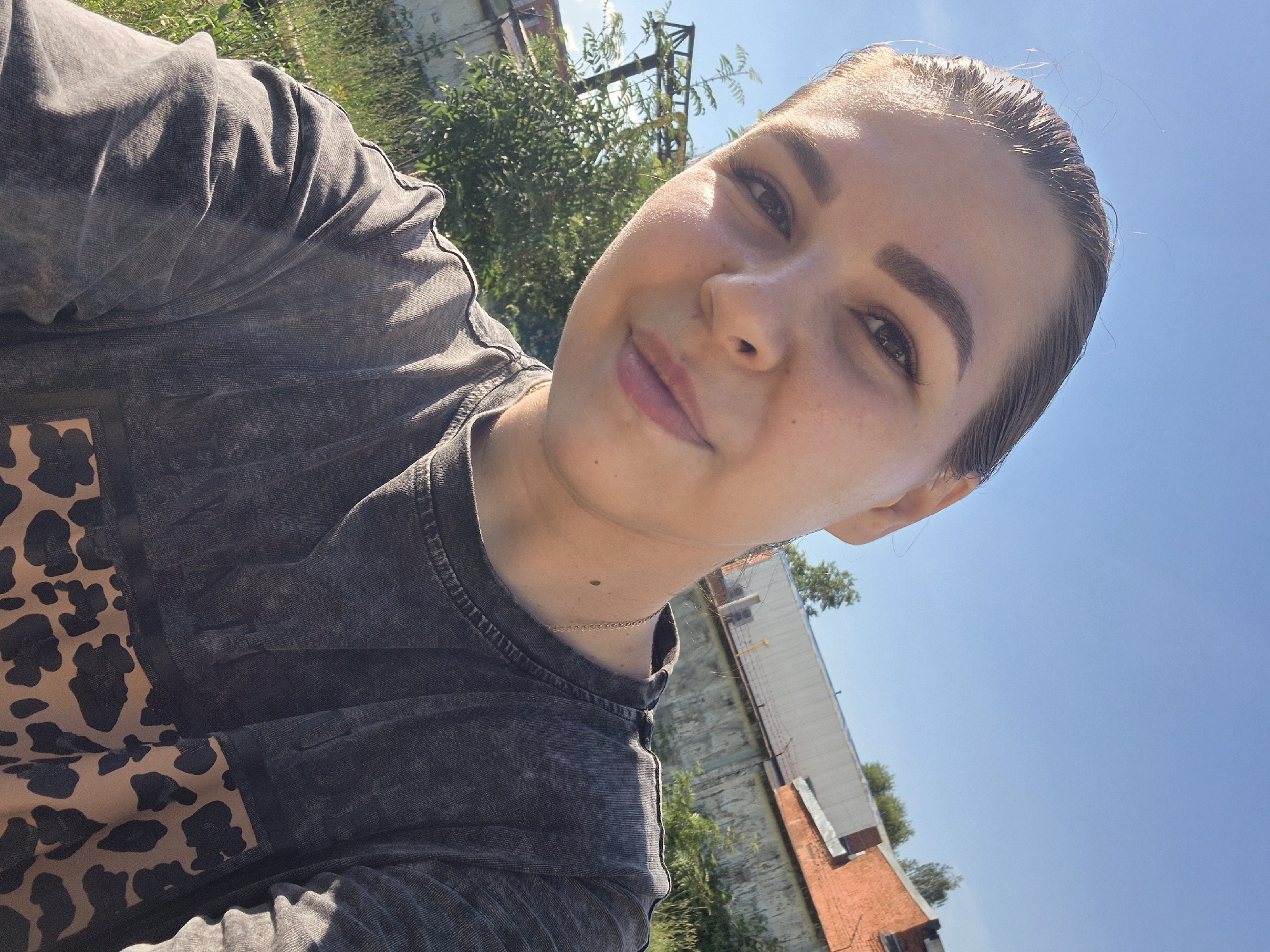 A young woman with her hair pulled back is taking a selfie outdoors. She is wearing a dark gray t-shirt with a leopard print panel on the lower right side. She has light-colored skin and is smiling slightly, with her head tilted to the side. The background shows a bright blue sky, green trees, and a brick building with a white section. There are also some industrial structures, including what appears to be a yellow crane, visible behind the trees. The scene suggests a sunny day in an urban or industrial area of Chaikovsky, Russia.