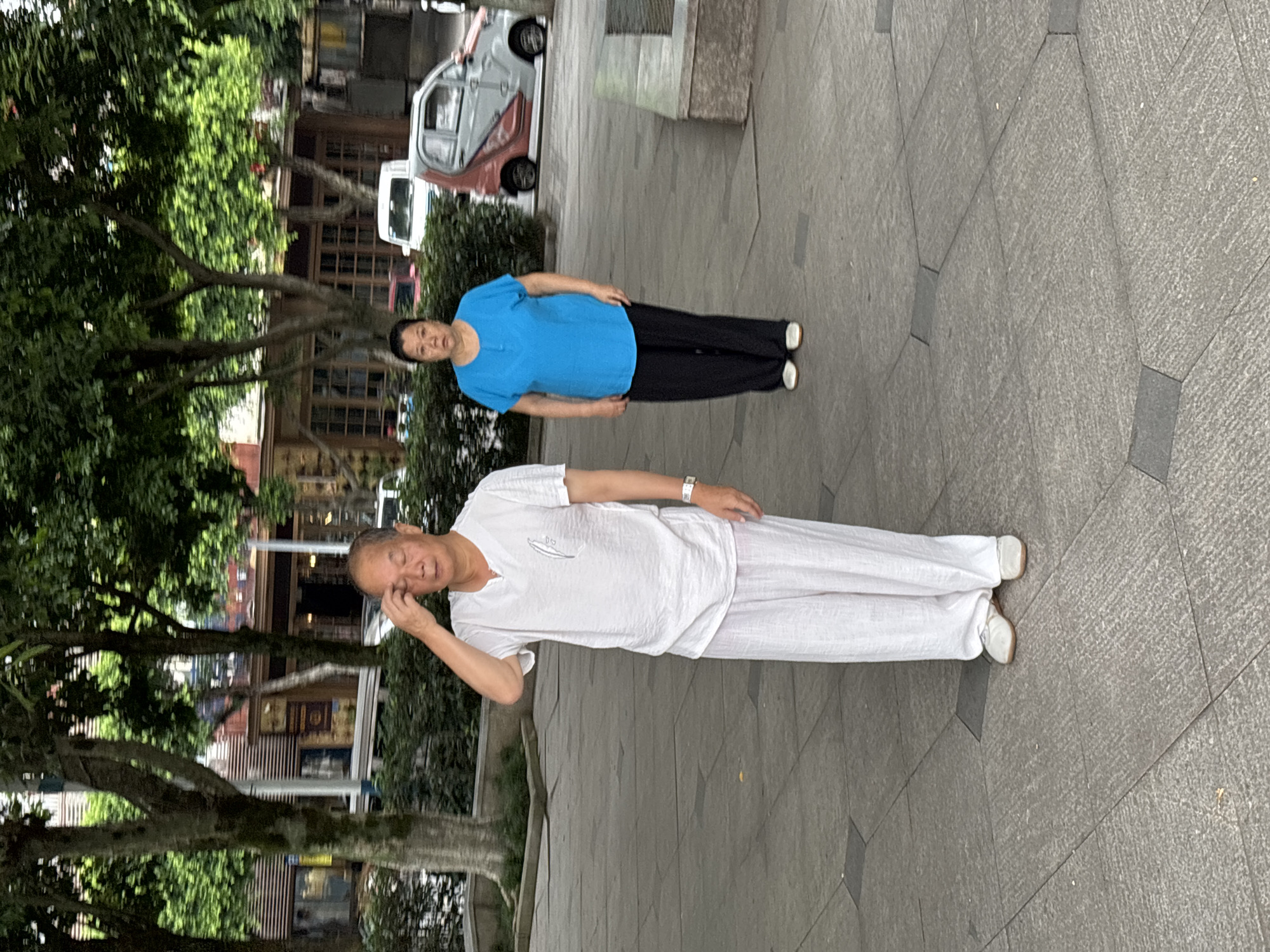 In the city of Jiangjin, China, on a paved outdoor area, an elderly Asian man stands in the foreground, wearing a loose-fitting white short-sleeved shirt and loose white pants. He has his right hand on his forehead, eyes closed. Behind him and to his right, a middle-aged Asian woman stands, wearing a bright blue short-sleeved shirt and dark pants. Her expression is neutral. The background features trees, bushes, and buildings, suggesting an urban park or pedestrian area. A small, uniquely designed car with a pink and gray pattern is visible in the mid-ground, along with other vehicles and structures that indicate a street scene. The lighting suggests daytime.