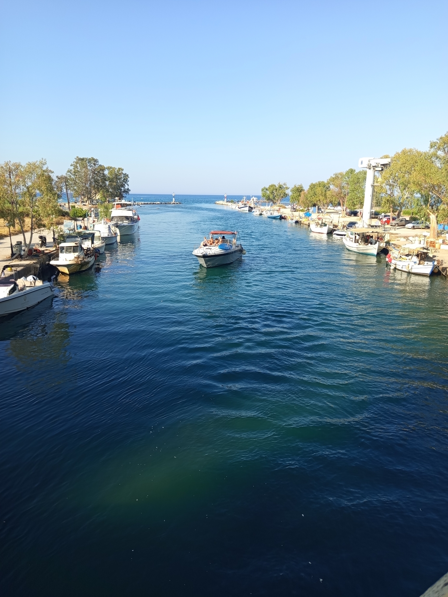 This is a wide-angle, eye-level outdoor shot of a clear blue sky above a channel of calm, dark blue water, likely in Georgioupoli, Greece, given the context. The channel is lined on both sides with moored boats and trees. On the left bank, there are several white and grey motorboats of various sizes, some appearing to be fishing vessels and others more recreational. A paved walkway runs along the bank, with streetlights and some parked cars visible behind the boats. On the right bank, there is a similar arrangement of moored boats, including some larger white vessels. A tall, white crane structure is prominent on this side, and more parked cars are visible behind the boats and trees. A few people are visible on the boats, one group lounging on the stern of a moving boat in the middle of the channel. The water is mostly a deep blue, with lighter turquoise hues near the edges and where sunlight penetrates. The distant horizon shows the sea meeting the sky, with two small lighthouses visible on either side of the channel entrance. The overall atmosphere is peaceful and sunny.