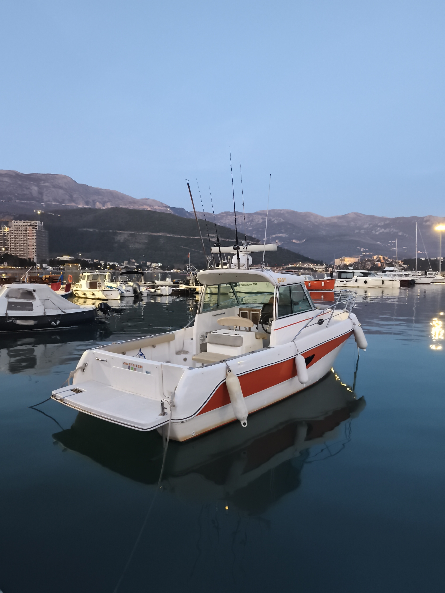 The image shows a white and orange motorboat docked in a marina in Budva, Montenegro. The boat is facing away from the camera and has fishing rods mounted on the roof. The boat's reflection is visible in the calm water. In the background, there are mountains, buildings, and other boats docked in the marina. The sky is a clear light blue. The time of day appears to be dusk or early evening.