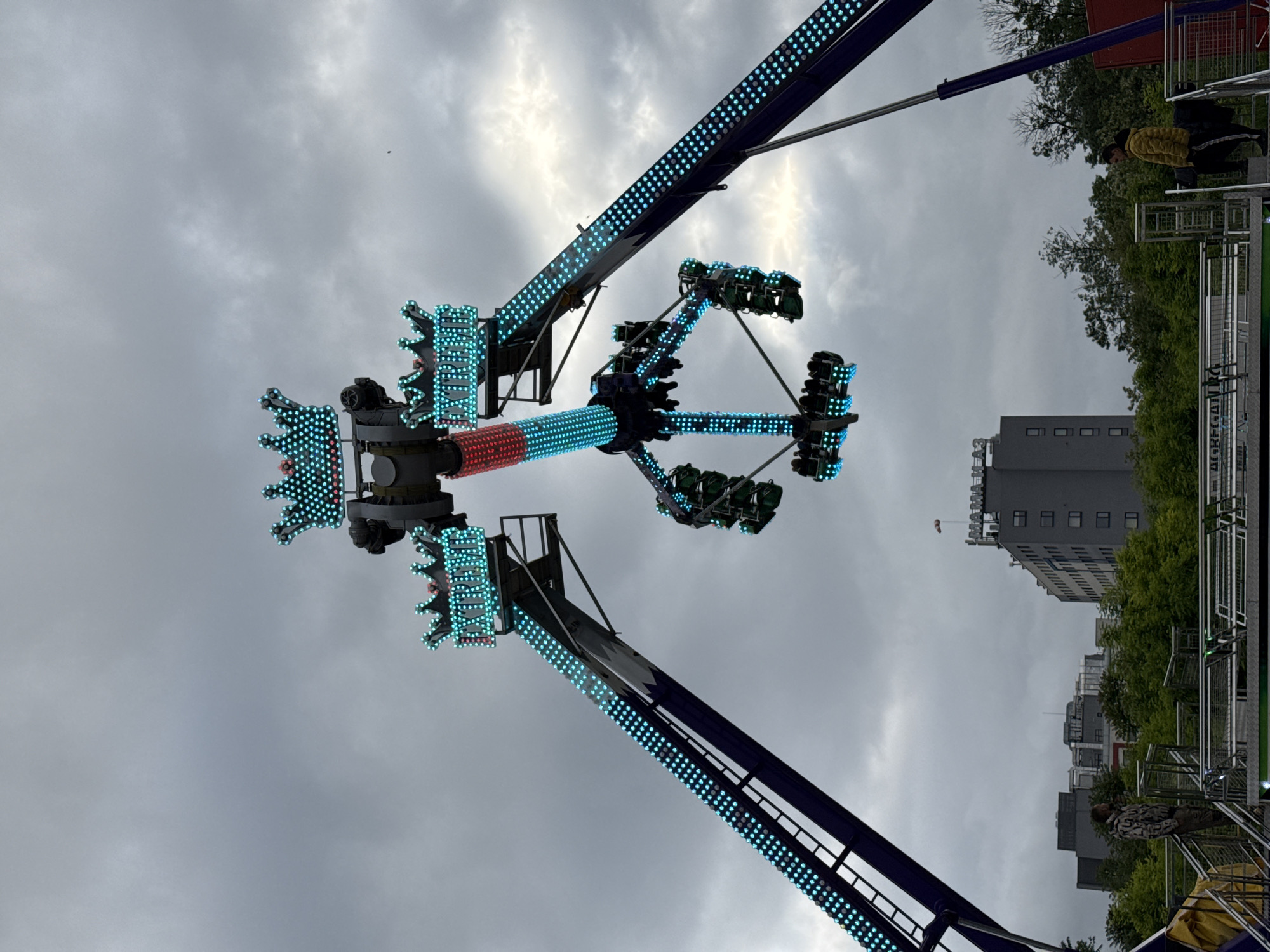 An outdoor photo displays a fairground ride, possibly a "sky dancer" or similar, with passenger carriages suspended from a central spinning arm. The ride is illuminated with blue and red LED lights. The sky is overcast and grey. A building and trees are in the background, and some people are visible on the right side, along with what seems to be ride fencing. It is believed that this image was captured in Pleven, Bulgaria.