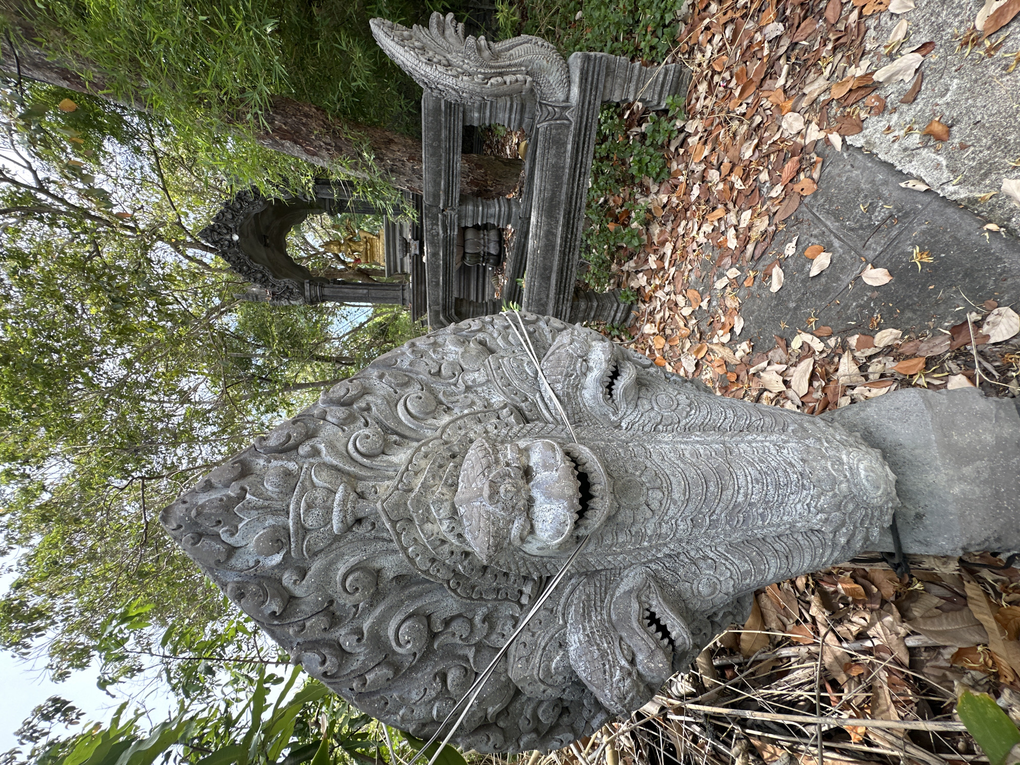 A weathered, multi-headed stone serpent sculpture stands in the foreground on a bed of fallen leaves. Behind it, a stone balustrade runs horizontally across the frame, leading to a small arched shrine containing a golden Buddha statue. The scene is set outdoors amidst dense green foliage and trees. The ground is covered in dry leaves and scattered patches of green vegetation. The sky is visible through the canopy, appearing bright.