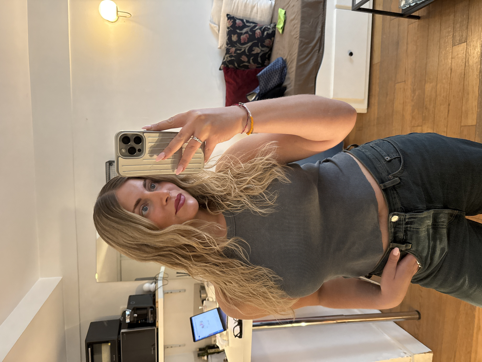 A young woman with blonde, wavy hair and blue eyes is taking a selfie in a room. She is wearing a grey ribbed crop top and dark wash jeans. Her left hand is in her front pocket. She is wearing a ring on her right index finger and a beaded bracelet on her right wrist. In the background, there is a bed with pillows, a shelf with kitchen appliances, a laptop, and a lamp on the wall. The room appears to be a studio apartment. There is no indication of the location being Paris, France.