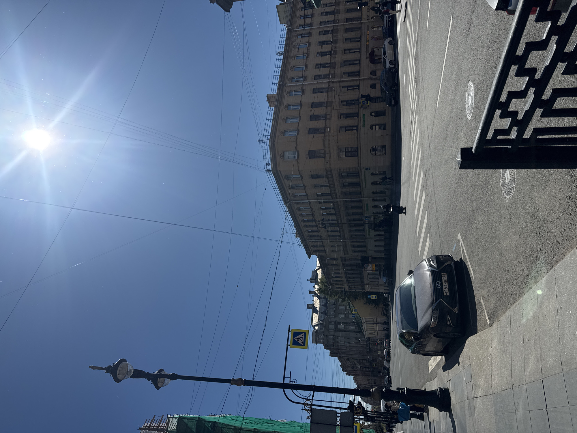 The image captures a street scene in Saint Petersburg, Russia, on a bright, sunny day. The sky is a clear, vibrant blue, with the sun shining intensely, creating lens flares and visible light rays. Overhead, a network of power lines crisscrosses the sky.
In the foreground, a dark-colored Lexus sedan is parked on the right side of the road, facing away from the camera, positioned over a white parking line. Its license plate reads "M151HB 198". To the left, a sidewalk with a tiled pattern is visible, bordered by a black lamppost with two lights. A black decorative metal fence is in the lower right corner of the frame.
The street itself is paved and shows clear lane markings and a pedestrian crossing. Several other cars are visible in the distance, driving or parked. To the left of the street, a building under construction is partially visible, covered in green netting.
The background features a row of multi-story buildings, characteristic of Saint Petersburg architecture, with many windows and ornate facades. A pedestrian crossing sign, a yellow square with a black pedestrian icon, is mounted on a pole. A few pedestrians are scattered along the sidewalk and crossing the street. One person is seen riding a motorcycle in the middle ground. The overall impression is a bustling urban environment under a clear, sunny sky.