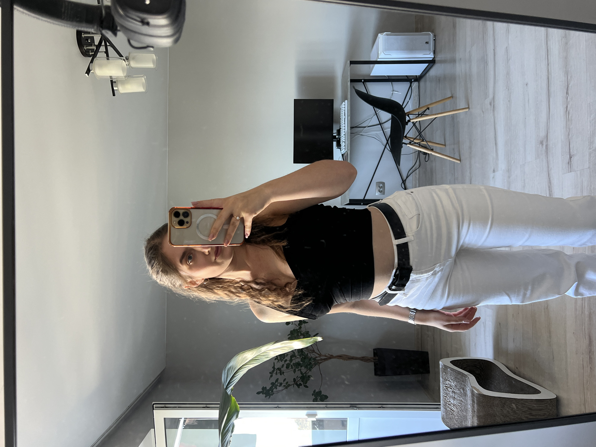 The image is a mirror selfie taken by a young woman with light brown hair, wearing a black crop top and white jeans. She is holding a smartphone in her right hand and appears to be in an indoor setting. The background shows a room with a desk, a computer monitor, a chair, and a plant. There are also reflections in the mirror of a chandelier and a window. The image is likely taken in Ternopil, Ukraine.