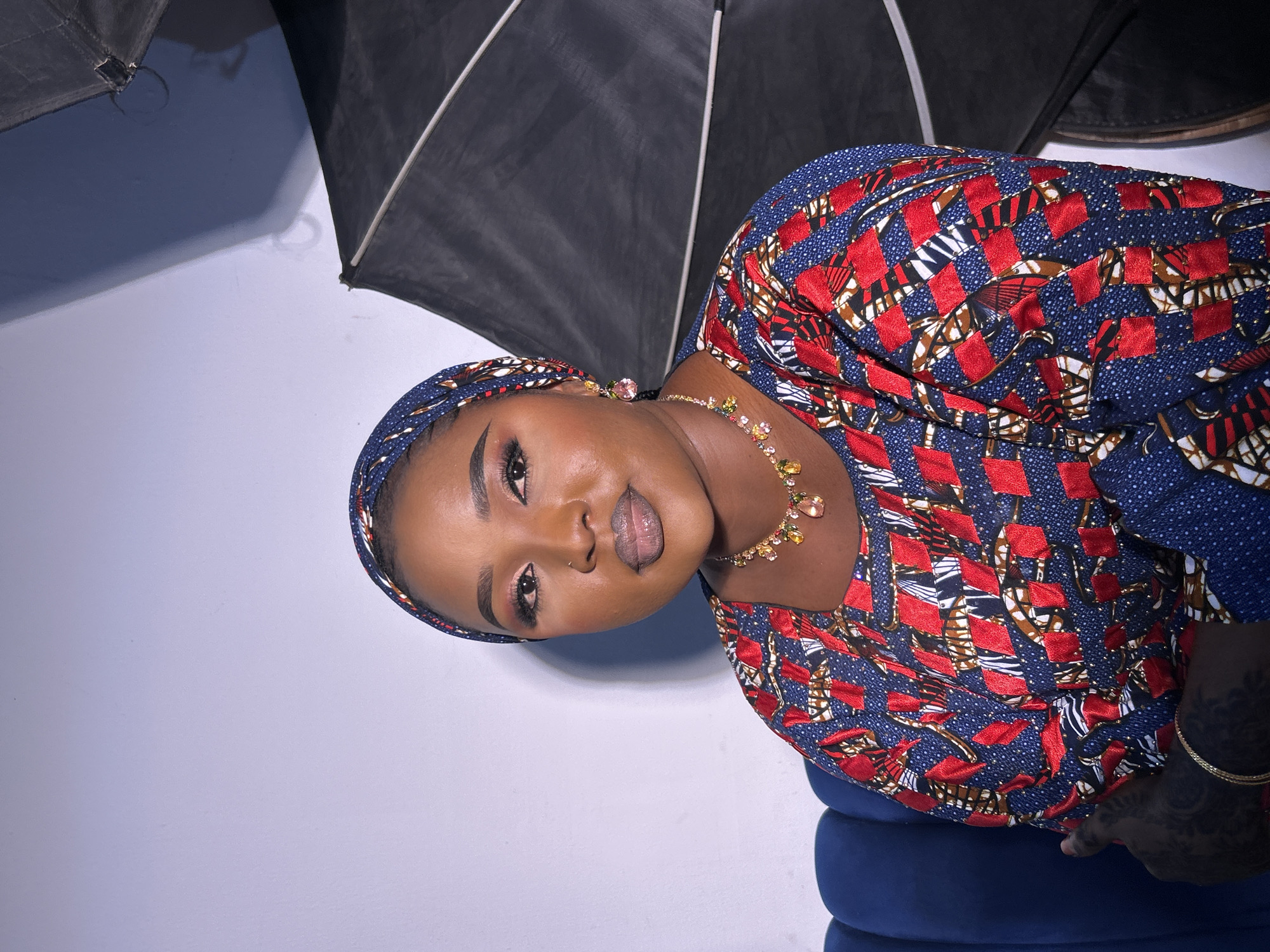 A woman is seated in front of a white wall, wearing a blue headscarf and a dress with a red, white, and brown pattern. A large, black lighting umbrella is positioned behind her. She wears a multicolored jeweled necklace and has visible makeup. The location is not specified in the image but is requested as the city of Jos, Nigeria.