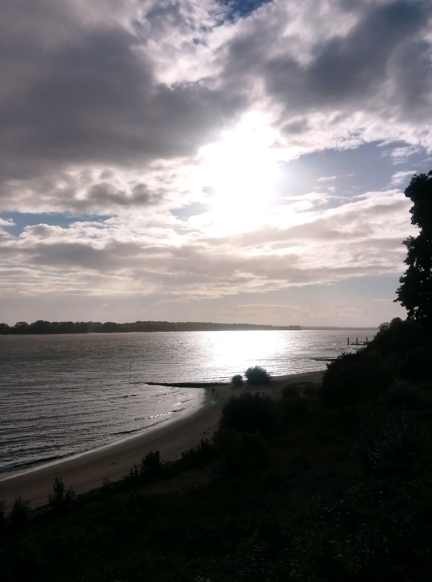 The image depicts a view of the Elbe river and its shoreline in Wedel, Germany, under a cloudy sky. The sun is partially obscured by clouds, creating a bright, hazy glow on the water's surface. The shoreline is composed of sand and vegetation, with trees and bushes visible along the right side of the frame. A wooden pier with a few small structures extends into the water. The overall atmosphere is serene, with a sense of natural beauty and tranquility. The image appears to have been taken from a slightly elevated position, looking down towards the river and the opposite bank, which is lined with trees.