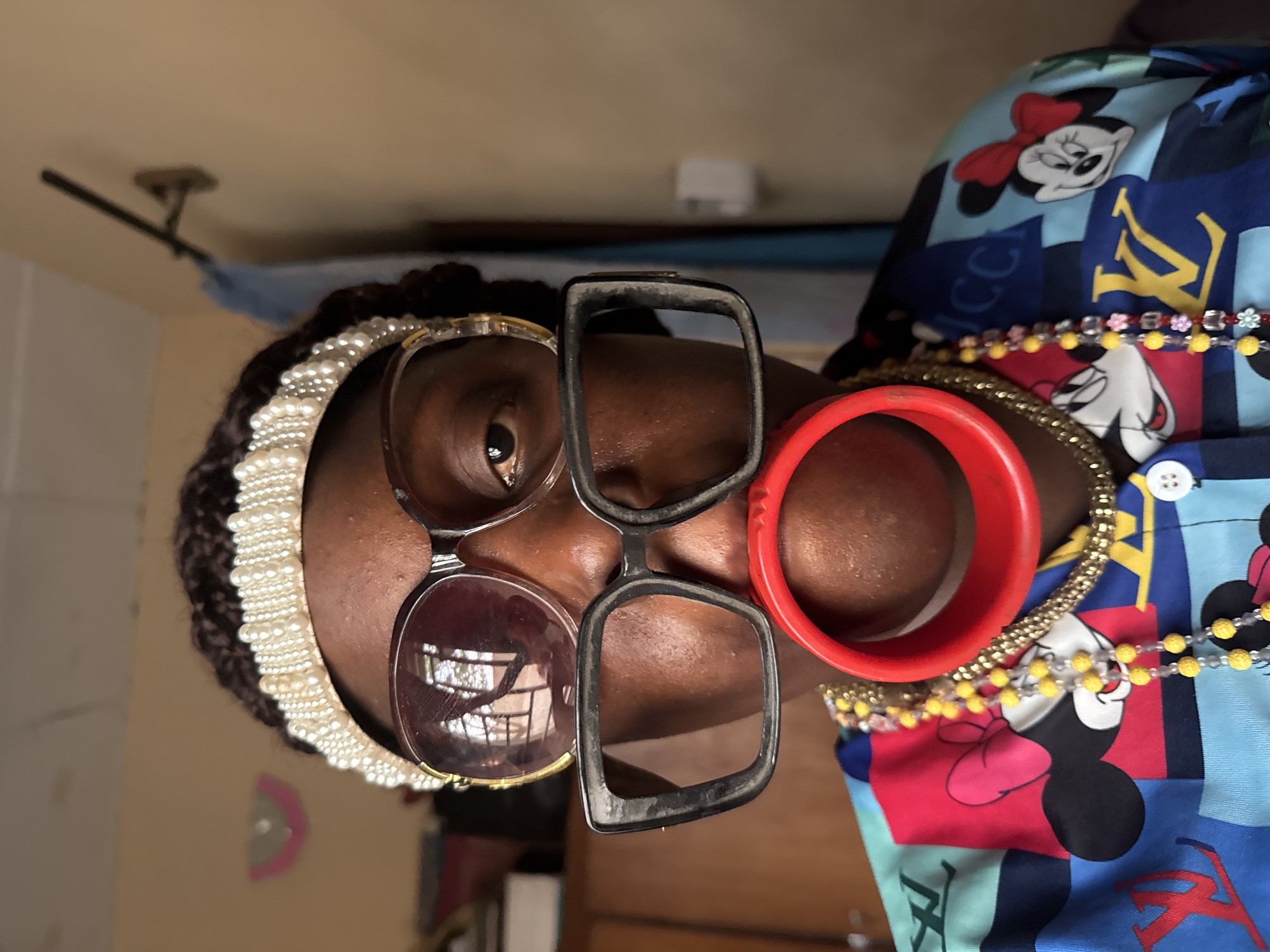 A medium close-up, slightly off-center, portrait-style photograph shows a dark-skinned person with a variety of accessories on their face and head. The photograph is rotated 90 degrees counter-clockwise.
The person wears a pearl-studded headband. Two pairs of eyeglasses, one tinted with round frames and the other with clear rectangular frames, are stacked in front of their face. A red, circular bangle sits next to their cheek. Multiple beaded necklaces, one gold and one with yellow and clear beads, adorn their neck. They wear a collared shirt printed with a pattern of cartoon characters, likely Mickey and Minnie Mouse, overlaid on a background of designer logos.
The background shows a light-yellow wall on the left, with a blurry, indiscernible object in the lower left corner. A white molding and blue accent wall are visible above. A metal rod hangs from the ceiling. The environment appears to be indoors.
The location cannot be determined, as the city of Madangala, Nigeria, is not depicted.