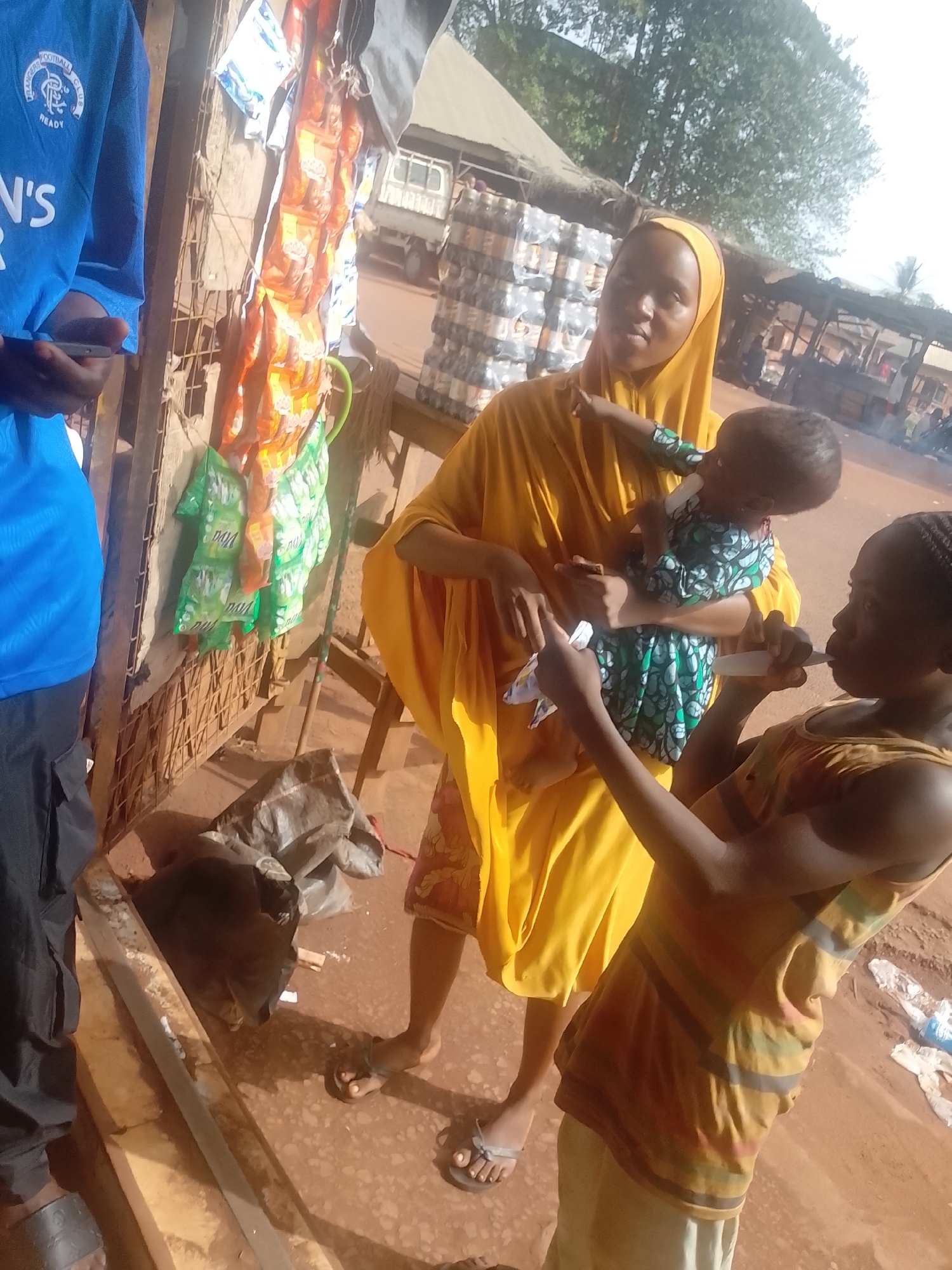 The image shows a scene in Ejule, Nigeria. A woman in a yellow headscarf and robe holds a baby wearing a green patterned outfit. A young person in a striped shirt stands beside them, holding a small white object to their mouth. A person in a blue Rangers football shirt stands to the left of the frame, partially obscured. Behind them is a weathered wooden structure with hanging merchandise. A truck and stacks of water bottles are visible in the background, indicating a possible marketplace or commercial area. The ground is reddish-brown.