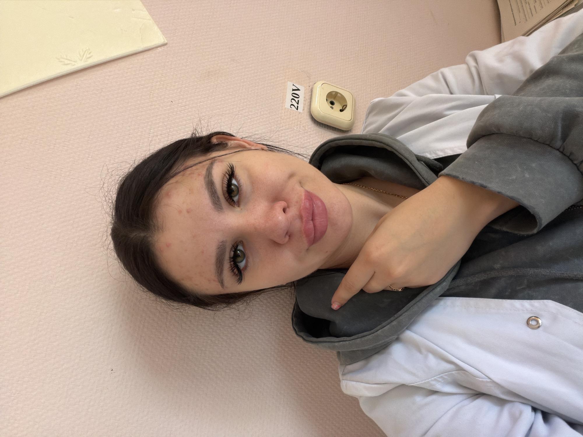 A young woman with dark hair and green eyes, exhibiting acne on her forehead and cheeks, is pictured in a close-up shot. She is wearing a white lab coat over a grey hoodie, and a gold necklace. Her right hand, adorned with a ring, is raised and pointing to a grey pillow held against her chest. To the right of the woman, on a pink textured wall, is a beige electrical outlet labeled "220V". The background also shows a portion of a pale yellow rectangular object with a faint embossed pattern. The scene appears to be indoors, possibly in a room with pale pink walls. The image composition suggests a selfie taken by the woman.