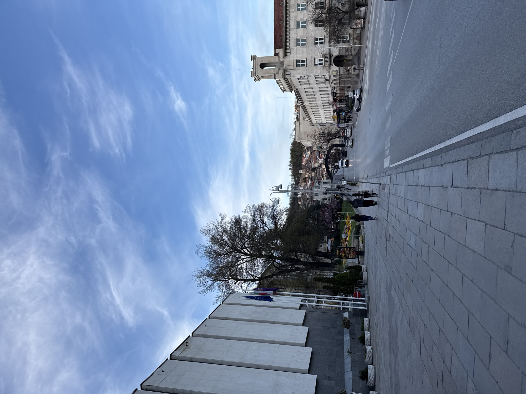 The image shows a street scene in Veliko Tarnovo, Bulgaria, under a partly cloudy blue sky. The foreground features a modern building with a flag display. Adjacent is a tree. The sidewalk leads to a road with moving vehicles, including cars and buses. In the background, there is a hill. The buildings appear to be in the downtown area. People are walking on the sidewalk, visible as silhouettes.