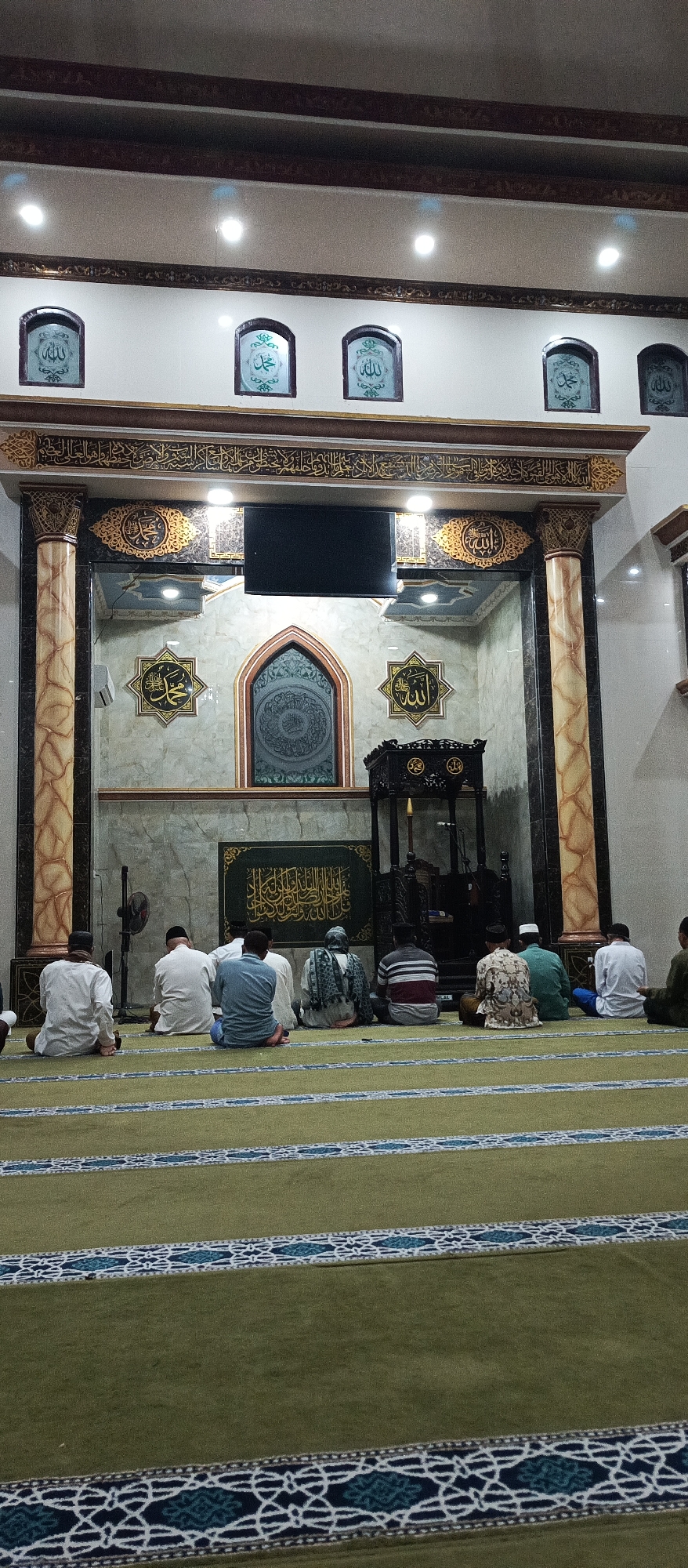 In the city of Arjasa, Indonesia, this image captures the interior of a mosque. Several individuals, likely congregants, are seated on a green carpeted floor, facing the front of the mosque where a prayer niche (mihrab) is visible. The mihrab is adorned with intricate Islamic calligraphy and has a decorative arched doorway. Above the mihrab, a television screen is mounted on the wall. The walls of the mosque are decorated with ornate Islamic geometric patterns and calligraphy. Fluted columns flank the central prayer area. The lighting appears to be from overhead fixtures. The overall atmosphere suggests a peaceful and devotional setting.