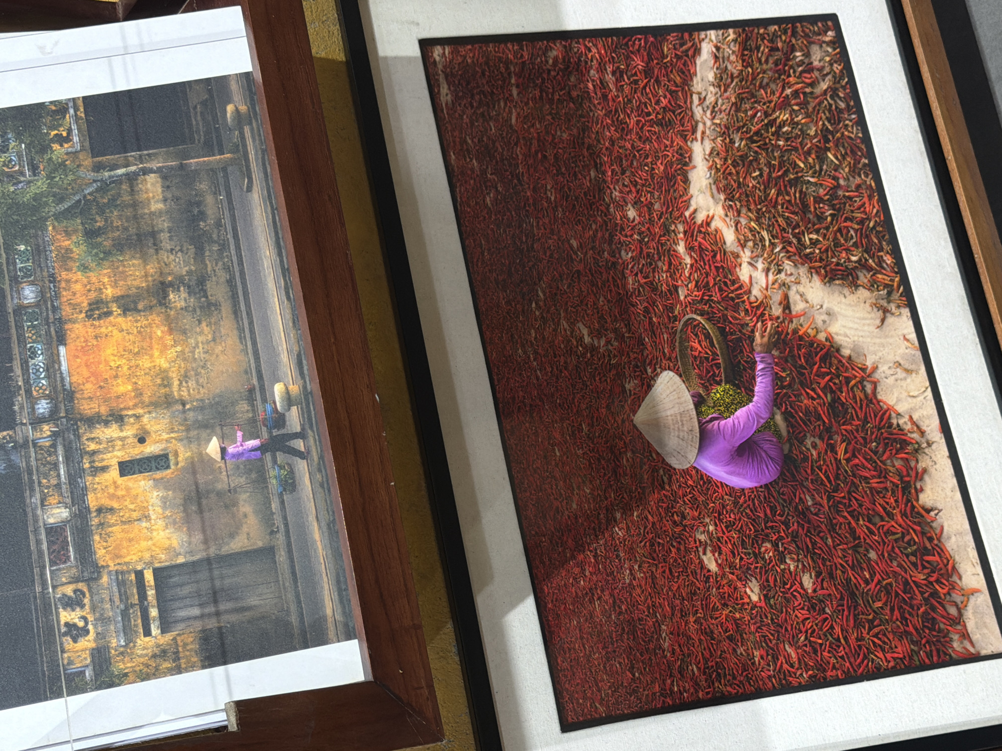 The media file displays two framed photographs, positioned at an angle. The most prominent image, on the right, captures an overhead perspective of a person surrounded by a vast spread of bright red chili peppers laid out on light-colored ground, likely for drying. The individual wears a striking purple tunic and a traditional Vietnamese conical hat (nón lá), and has a woven basket nearby. The scene is well-lit, creating clear shadows from the person and hat, suggesting daytime.
To the left, a second framed photograph is partially visible. This image depicts a street scene with an aged building characterized by earthy yellow and brown tones. Two figures wearing conical hats are present, one of whom is dressed in purple and carrying a shoulder pole with baskets, a common sight in Vietnamese markets or rural areas. Both photographs showcase elements of Vietnamese culture and daily life, consistent with the provided location context of Hội An Ward. The right photograph features a dark frame with a white mat, while the left one has a brown wooden frame. There is no visible text in either image or on their frames.