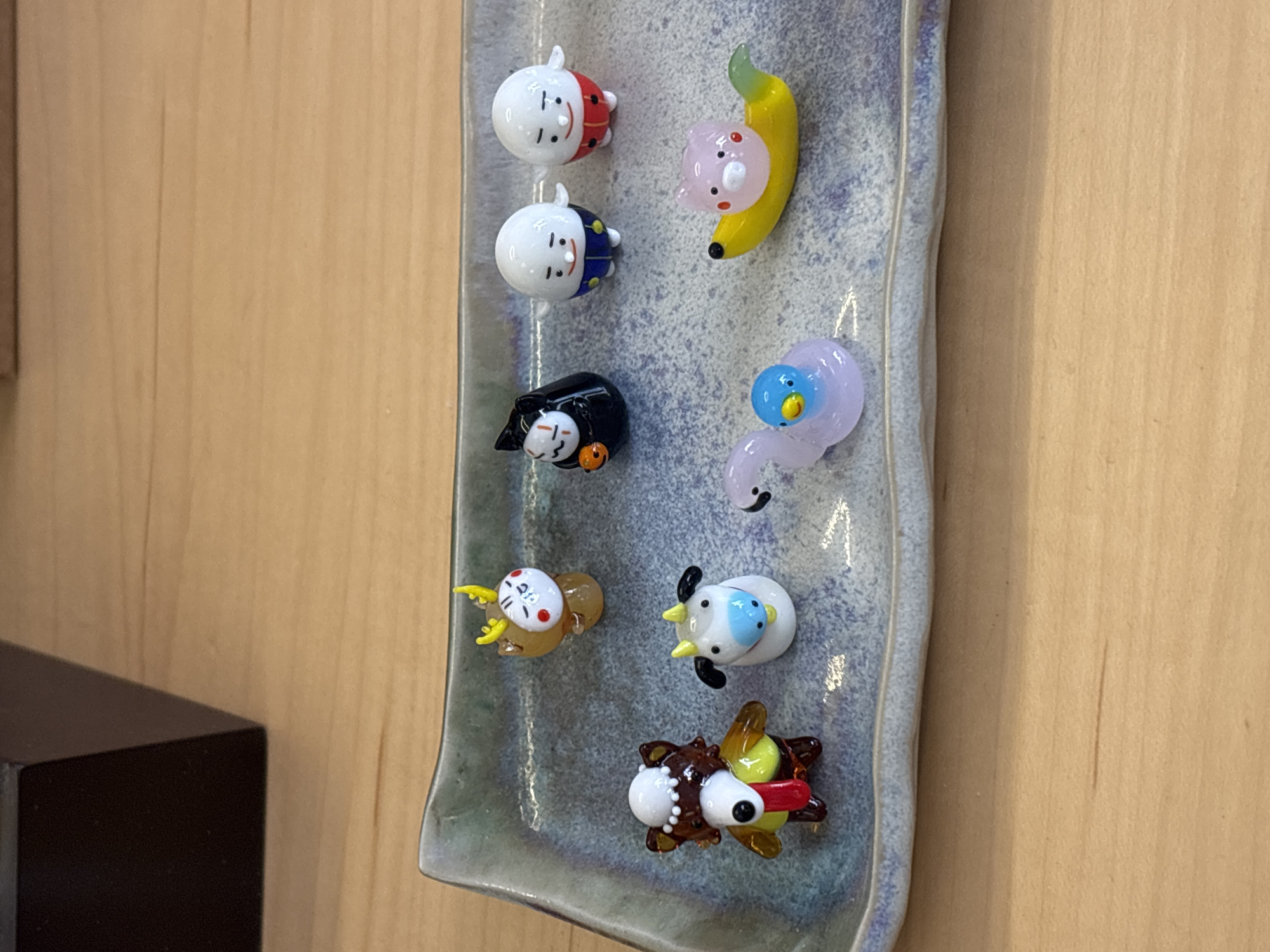 The image shows a display of small, glass figurines arranged on a rectangular, ceramic tray with a textured, gray-blue glaze and wavy edges. The tray is mounted on a wall with a light-colored, wood-grain surface. From top to bottom, the figurines depict whimsical characters: two round-headed figures with painted faces and red and blue caps respectively, a pink pig riding a yellow banana, a black cat with a ghost face and orange detail, a small brown and yellow bird-like creature with yellow wings, a white animal with a blue spot, and a brown bug-like creature with red and yellow features. The scene appears to be indoors, likely in a shop or gallery setting. The location in Madrid, Spain is not discernible from the image.
