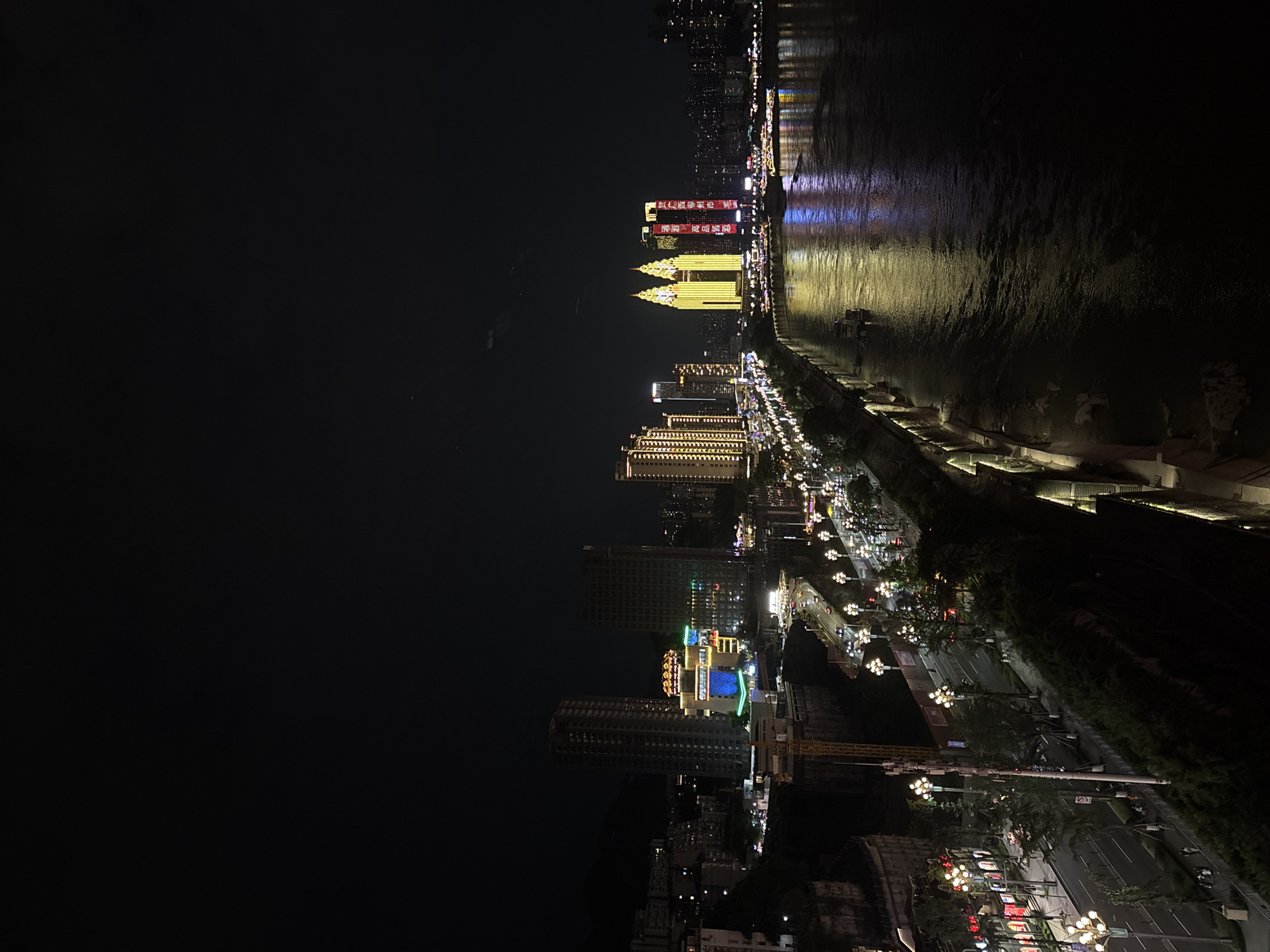 An aerial night view captures a bustling cityscape in Yuzhong District, China, illuminated by a vibrant display of lights. A wide river dominates the foreground, its surface reflecting the colorful glow of the city and passing boats. Along the riverbank, a well-lit promenade features decorative lampposts and lush greenery. Adjacent to the promenade, a multi-lane road carries vehicular traffic, with cars and streetlights creating streaks of light. Buildings of varying heights and architectural styles line the riverfront, some adorned with illuminated signs and decorative lighting, showcasing a dynamic urban landscape. In the distance, towering skyscrapers pierce the dark night sky, their windows a mosaic of light. The overall impression is one of a lively and active metropolis at night, with the river serving as a central artery reflecting the city's energetic pulse.