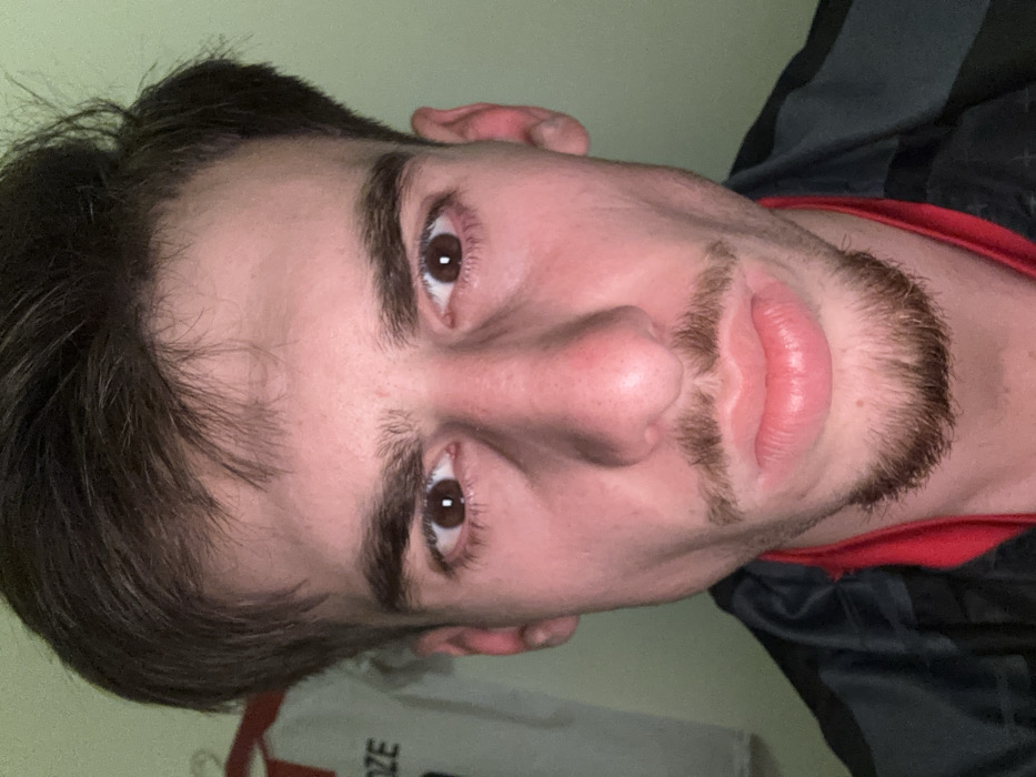 A close-up headshot depicts a young man with light skin, brown hair, and brown eyes. He has a dark mustache and goatee. The image is rotated, so he is positioned sideways. He is wearing a red shirt with a black and gray jacket. The background appears to be a dimly lit room with a white object partially visible near the bottom left corner.