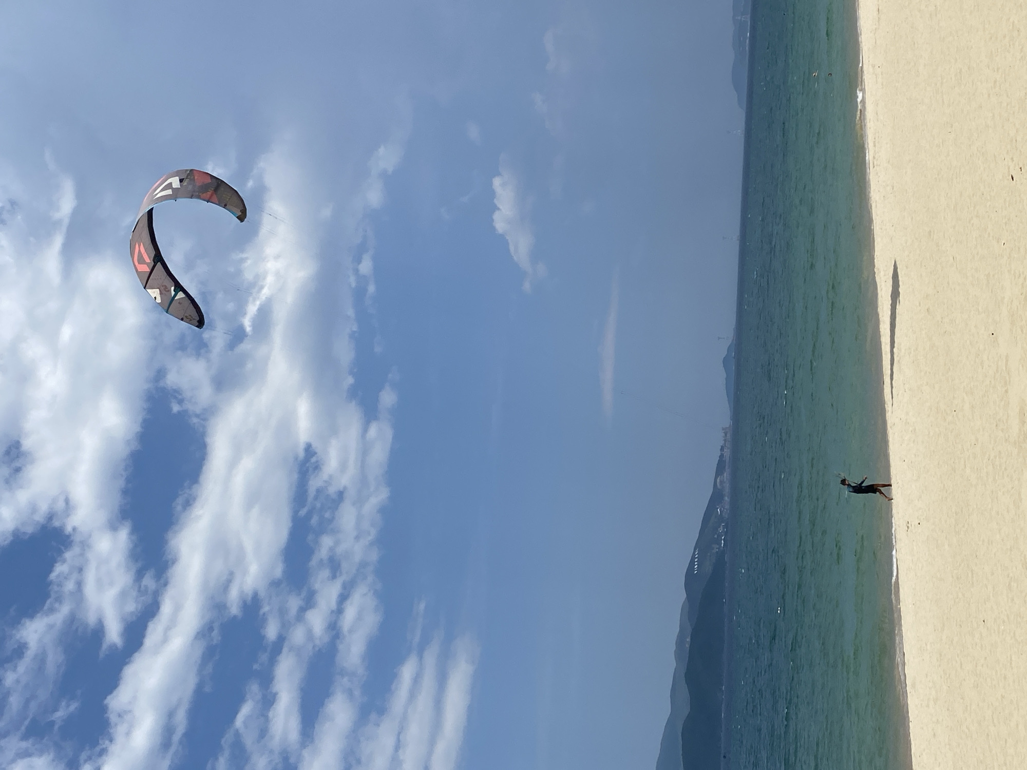 The image shows a sunny day at a beach in Nha Trang, Vietnam. A large, colorful kite is soaring in the blue sky, which is dotted with white, puffy clouds. In the middle ground, the turquoise ocean meets a sandy beach. A lone person, dressed in dark shorts and a dark top, is walking along the shoreline, possibly carrying a kiteboarding kite setup. In the background, a range of lush green hills forms the coastline. The overall scene depicts a leisurely day at the beach, with the kiteboarding activity adding a dynamic element to the peaceful landscape.
