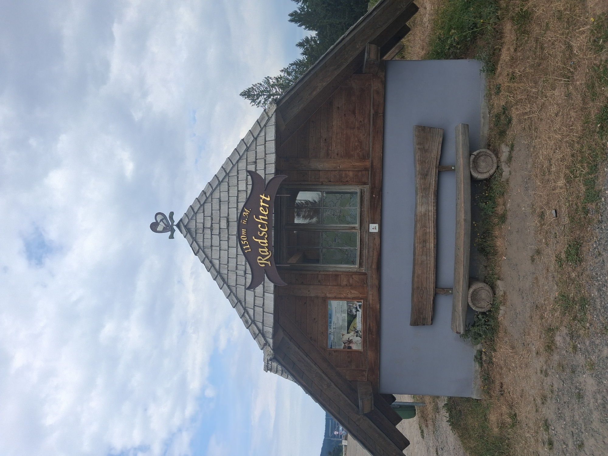 A rustic wooden and stucco building with a shingled roof stands under a cloudy sky. A sign above a window reads "1150m ü.M. Radschert". A wooden bench is positioned in front of the building. The building appears to be located in a natural, possibly mountainous, environment with trees in the background and dry grass in the foreground. The image is a full-shot photograph of the building.
