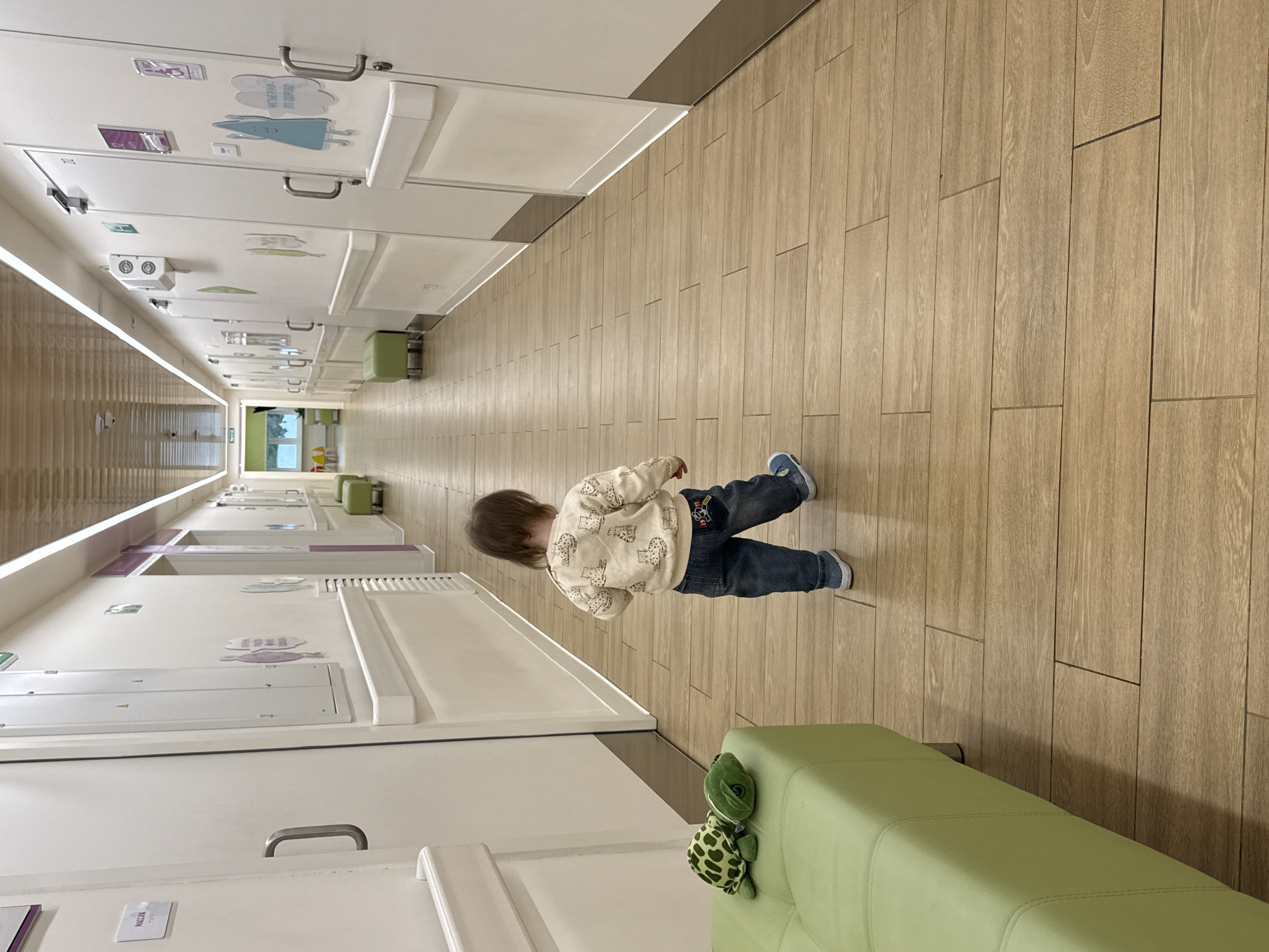 A young child, seen from behind, walks down a brightly lit hallway in what appears to be a modern children's facility. The child has brown hair and is wearing a cream-colored long-sleeved top with a repeating animal pattern, blue jeans, and blue sneakers. They are positioned on the right side of the wooden-planked floor, heading away from the viewer.
The hallway is characterized by a long, light-colored wooden floor. On both sides, white wall panels or cabinet units line the space, extending into the distance. These panels feature various decorative stickers or cutouts, such as a blue figure, purple shapes, and cloud-like designs, suggesting a child-friendly environment. Some panels have metal handles, indicating they are functional doors or compartments.
Along the right wall, closer to the foreground, a light green upholstered bench is visible, and perched on it is a green plush turtle toy with a patterned shell. Further down the hallway, other similar green benches can be seen. The ceiling is equipped with linear lighting fixtures that illuminate the space. At the far end of the hallway, a large window allows natural light to enter, revealing some greenery outside.
The scene depicts a calm daytime setting. One visible text label on a white panel reads "MACCON." Other decorative elements and small signs are present on the walls, but their text is not clearly legible. The overall impression is a clean, organized, and welcoming environment, likely a medical clinic or a children's learning center in Moscow, Russia.