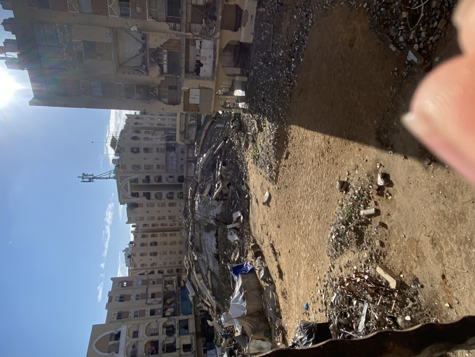 This image captures a wide view of a severely damaged urban area in Khan Yunis, Palestinian Territory, under a clear, bright blue sky with direct sunlight causing lens flare from the upper right. The foreground shows an uneven expanse of sandy earth and scattered rubble, with a blurred human finger partially obscuring the bottom right corner and a dark, indistinct object on the lower left edge.
The midground is dominated by a massive mound of building debris, consisting of broken concrete, twisted rebar, and other structural wreckage, indicating a completely collapsed structure. To the left of this central rubble pile, several makeshift shelters constructed from white and blue tarpaulins and fabric are erected on the ground. A single individual wearing a blue garment is visible standing near these temporary shelters.
The background features numerous multi-story residential or commercial buildings, all exhibiting extensive damage. Many structures have missing walls, exposing interior rooms, shattered windows, and visible pockmarks on their facades. Some buildings display distinct architectural features, such as arched window frames. On the rooftops, communication infrastructure is visible, including a prominent cellular tower antenna and what appear to be solar panels on another building. Strong shadows are cast across the ground and parts of the remaining structures due to the high sun.